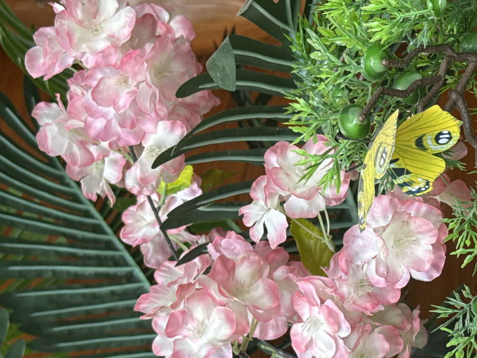 This image presents a close-up, frontal view of a decorative arrangement of artificial flora, located in the city of Furong, China. The primary subjects are clusters of artificial flowers, predominantly pink with white centers and edges, resembling hydrangeas or cherry blossoms, crafted from fabric. These flowers occupy the left and upper portions of the frame. Behind and interspersed with these flowers are large, dark green, segmented artificial leaves, suggestive of palm or fern fronds. In the lower right quadrant of the image, a mass of brighter green, finely textured artificial foliage is visible, featuring small, round, glossy green objects resembling berries. A gnarled, dark brown, artificial branch-like structure extends horizontally through this lighter green foliage. Affixed to the right side, resting on the lighter green foliage and brown branch, is a bright yellow artificial butterfly with distinct black markings on its wings. The background consists of a blurred, warm-toned surface, possibly wood or a wall. No human figures or active subjects are present; the scene depicts a static, ornamental display.
