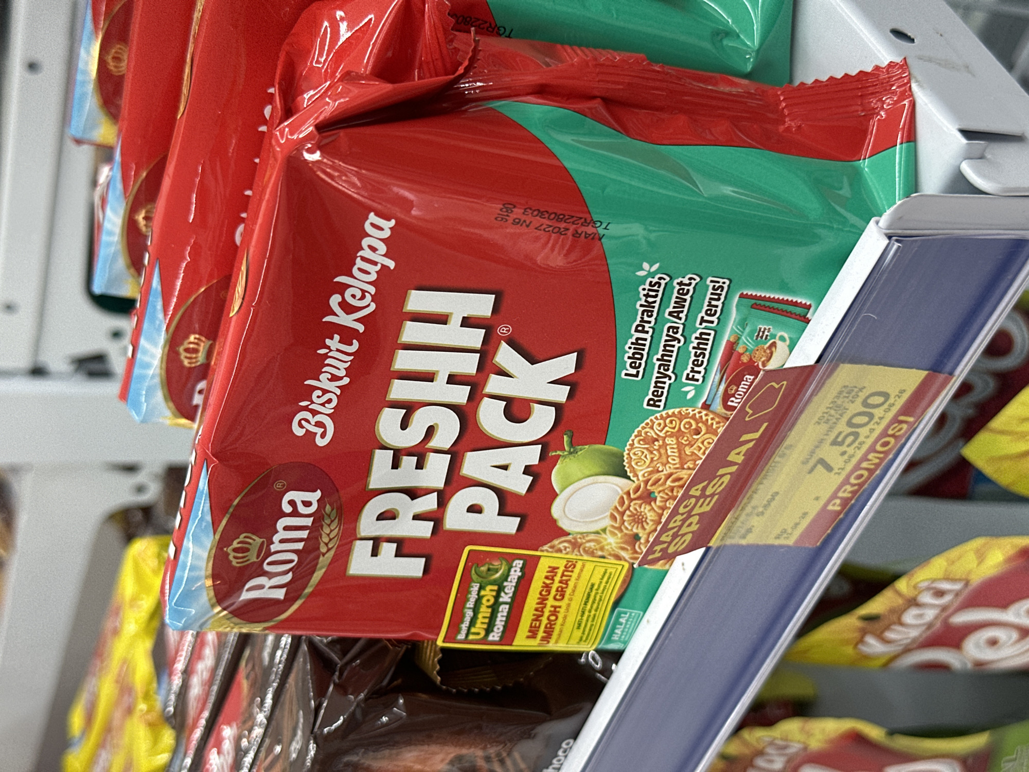 The image shows a close-up, eye-level, indoor shot of stacked red and green packaged biscuits on a shelf in what appears to be a retail store. The focus is on a red package with "Roma BiskuIt Kelapa FRESHH PACK" prominently displayed in white lettering. The packaging also features a golden crown logo, wheat stalks, and an illustration of a halved coconut. There are also yellow promotional tags on the shelf. The background is blurred, showing other packaged goods. The lighting is bright and even.