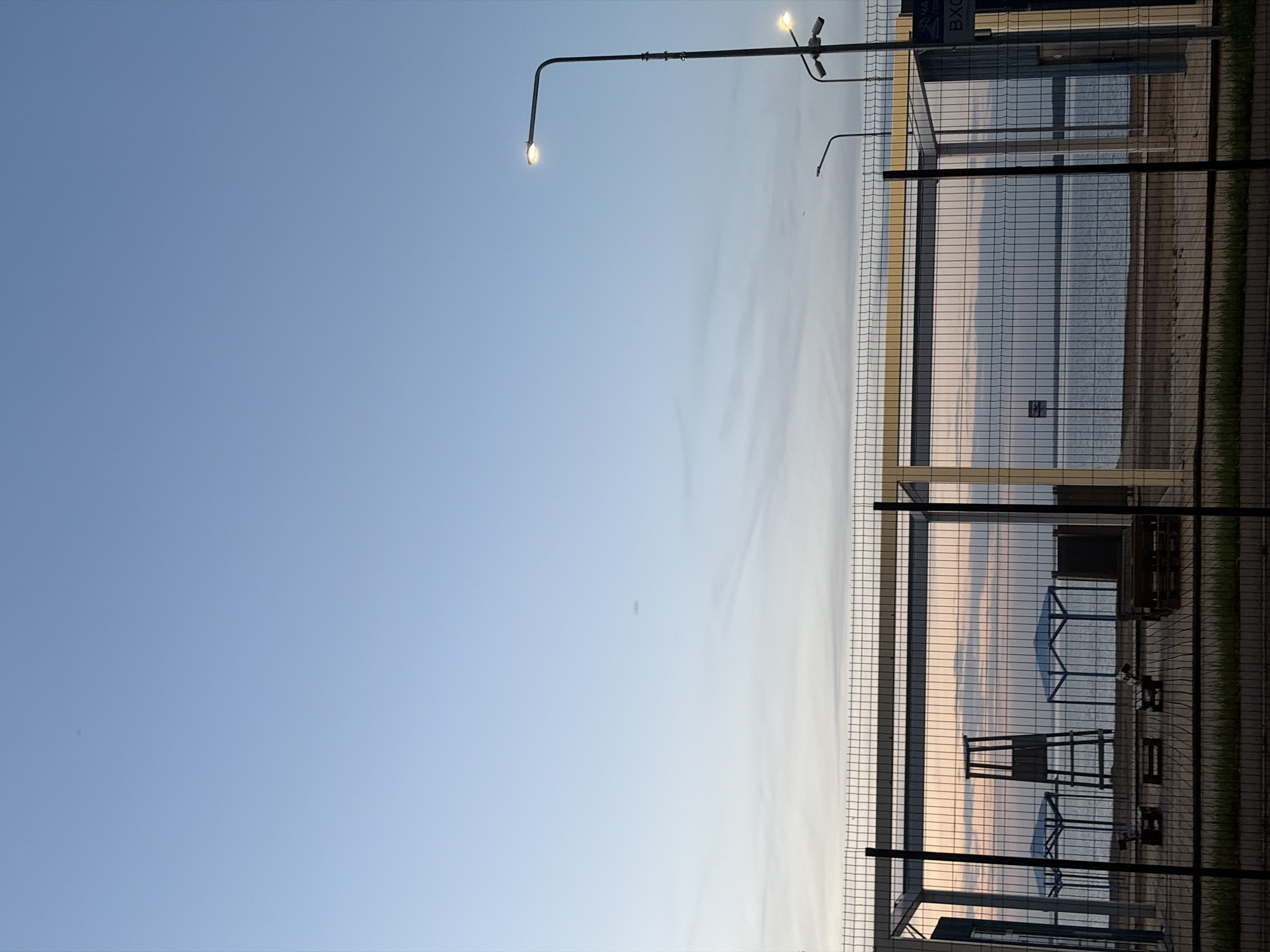 The image presents a vertical view of a cityscape in what appears to be Chaikovsky, Russia. The sky occupies the left side of the frame, showing a gradient from a darker blue at the top to a lighter, almost white hue towards the bottom. A street lamp with two illuminated bulbs is positioned on the upper left. On the right side, a metal mesh fence dominates the scene, providing a view through the fence of what appears to be a body of water. The water reflects the colors of the sunset sky, creating a visual contrast with the clear blue sky. Behind the fence, there are indistinct shapes that seem to be structures and possibly vegetation. The setting appears to be an urban environment, possibly near a shoreline.
