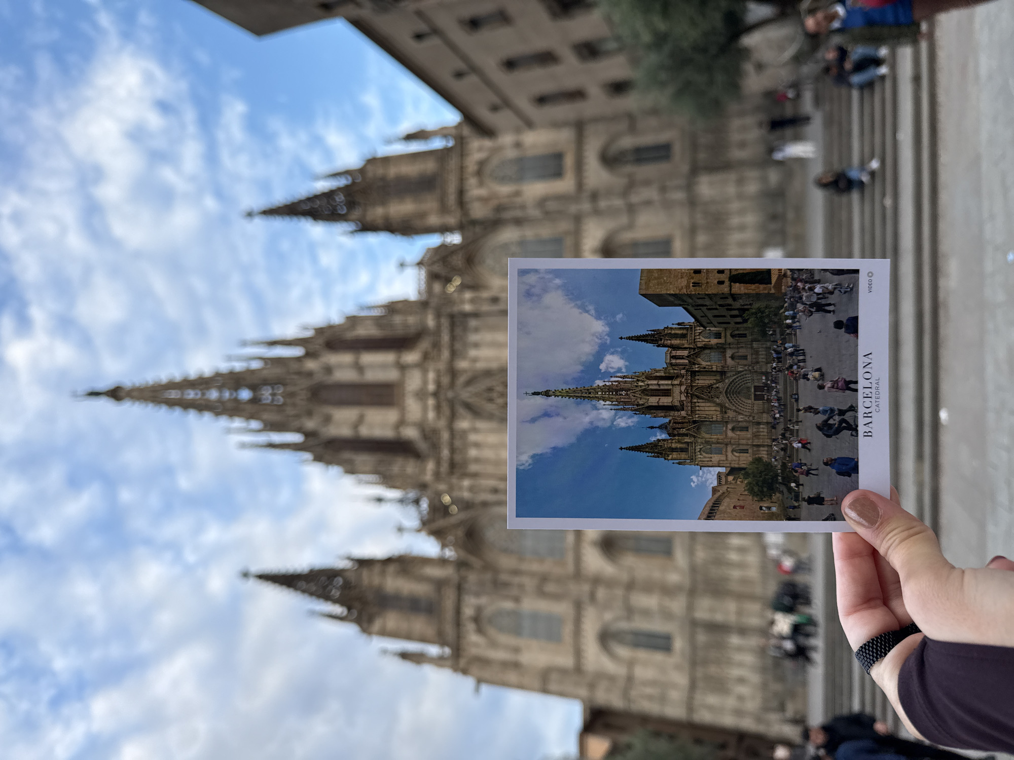 A person holds a postcard in front of Barcelona Cathedral in Barcelona, Spain. The postcard depicts the cathedral's facade and courtyard. The actual cathedral is visible in the background, with its spires reaching toward a blue, partly cloudy sky. People are present on the cathedral steps and plaza. The individual holding the postcard wears a black wristband.