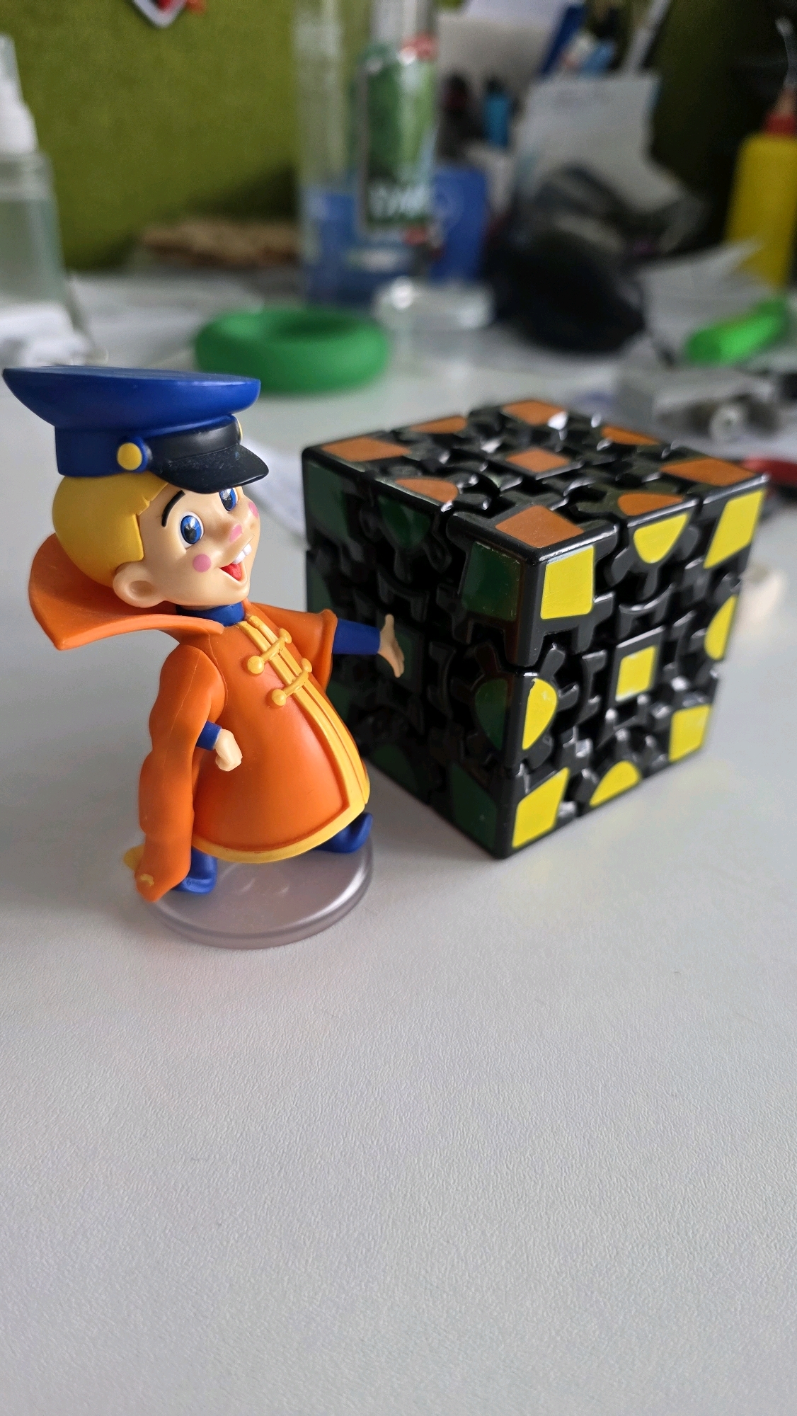 A colorful figurine of a young boy wearing a blue police hat and an orange coat with yellow trim stands to the left of a black gear puzzle cube with orange, yellow, and green accents. The figurine is standing on a clear circular base. The puzzle cube is positioned to the right of the figurine. The scene appears to be indoors on a white, textured surface. The background is blurred and shows hints of office supplies and bottles. This image does not provide any information about the city of Brest, Belarus.