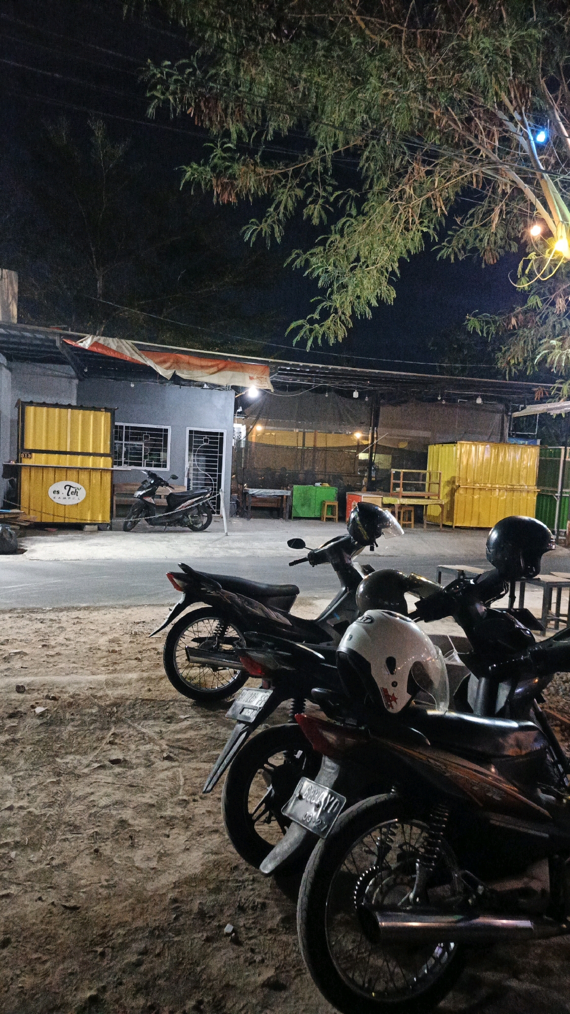 The image depicts a nighttime scene outside a small establishment in Pabelan, Indonesia. Several motorcycles are parked on a sandy or dirt ground in the foreground. Two helmets are placed on the seats of the motorcycles. In the background, there is a building with windows and doors, and yellow shipping containers are visible to the left. Lights are illuminating the area, and trees with foliage are overhead, with some string lights hanging from them. The overall impression is of a casual, outdoor gathering or dining area.
