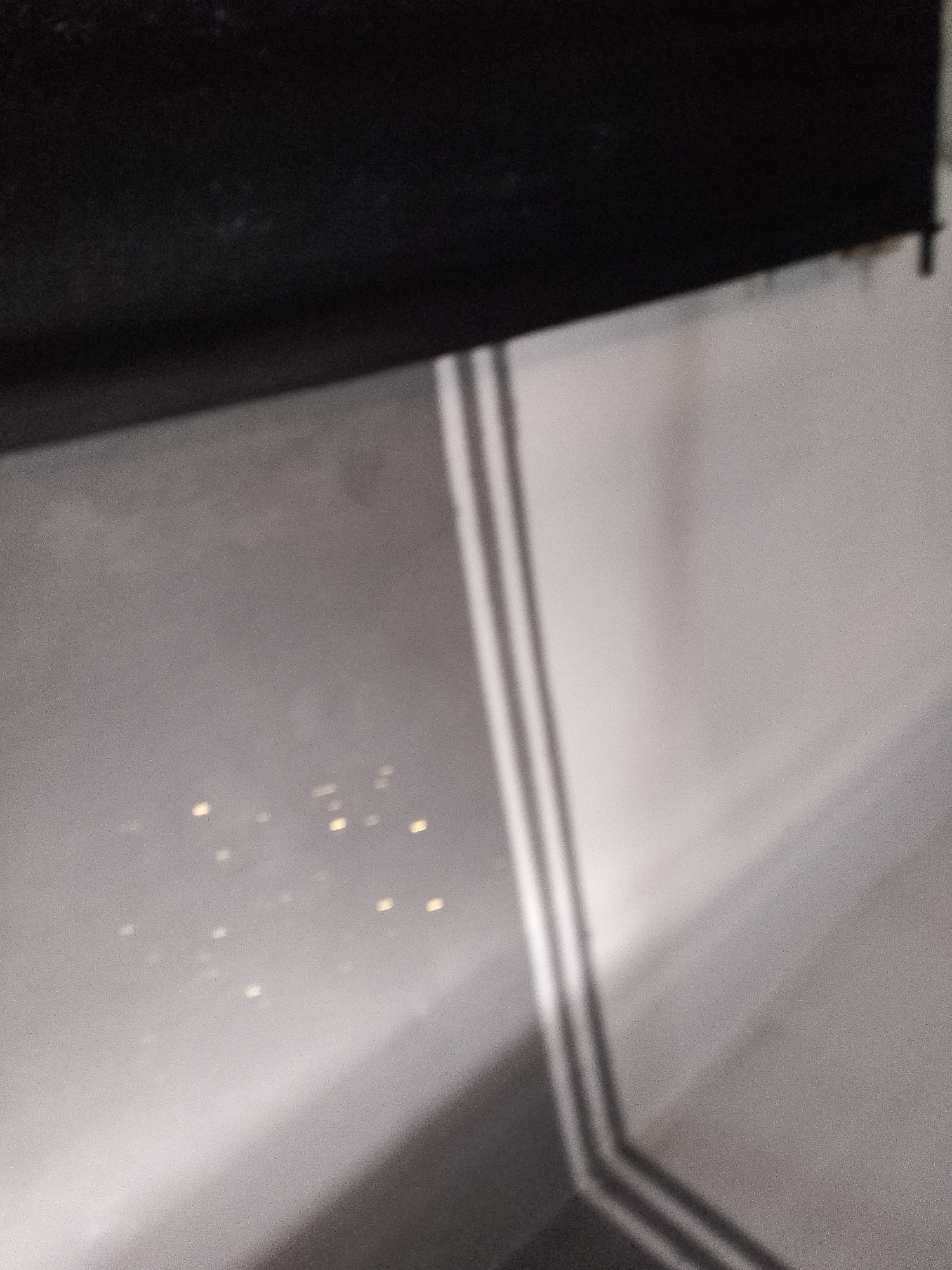 The image shows a close-up, low-light view of a window with a dark roller blind at the top. The window appears to be dirty or smudged, with several small, blurry yellow lights visible on the exterior, possibly from distant buildings. The reflections and light patterns on the window suggest it is nighttime. The overall scene is grainy and out of focus, making it difficult to discern specific details about the environment or location.
