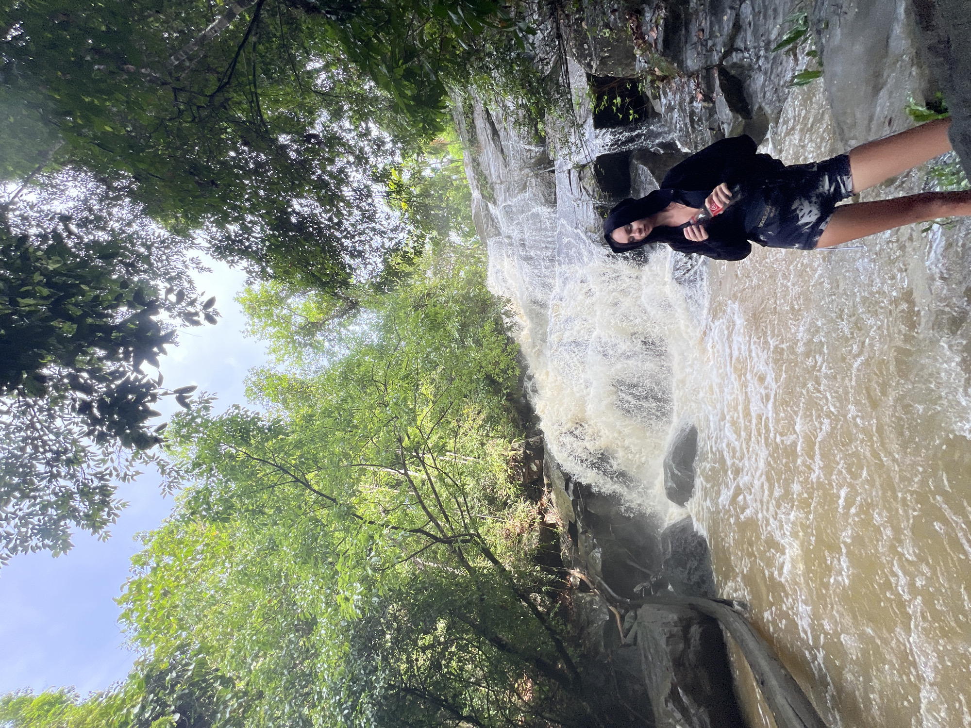 A brightly lit, vertically oriented photograph captures a person standing in the turbulent, brown water at the base of a powerful waterfall. The setting is a lush, natural environment, likely a tropical forest, identified as Siyambalagoda, Sri Lanka.
The main subject is a young person, possibly female, standing with their back to a rocky cliff from which the waterfall descends. They are wearing a black hooded top and dark-patterned shorts. In their right hand, they hold a plastic bottle with a prominent red label and white text, resembling a Coca-Cola bottle. Their expression appears calm or contemplative.
The waterfall itself is a significant feature, with white, churning water cascading over dark, rugged rock formations. The water in the pool at the base where the person stands is murky and brownish, indicating a strong flow that has stirred up sediment. Surrounding the waterfall and the rocky terrain is dense, vibrant green foliage and numerous trees, some of which frame the top and left side of the image. Through the gaps in the tree canopy, patches of bright, clear sky are visible, suggesting it is daytime. The overall impression is one of immersion in a powerful natural landscape.