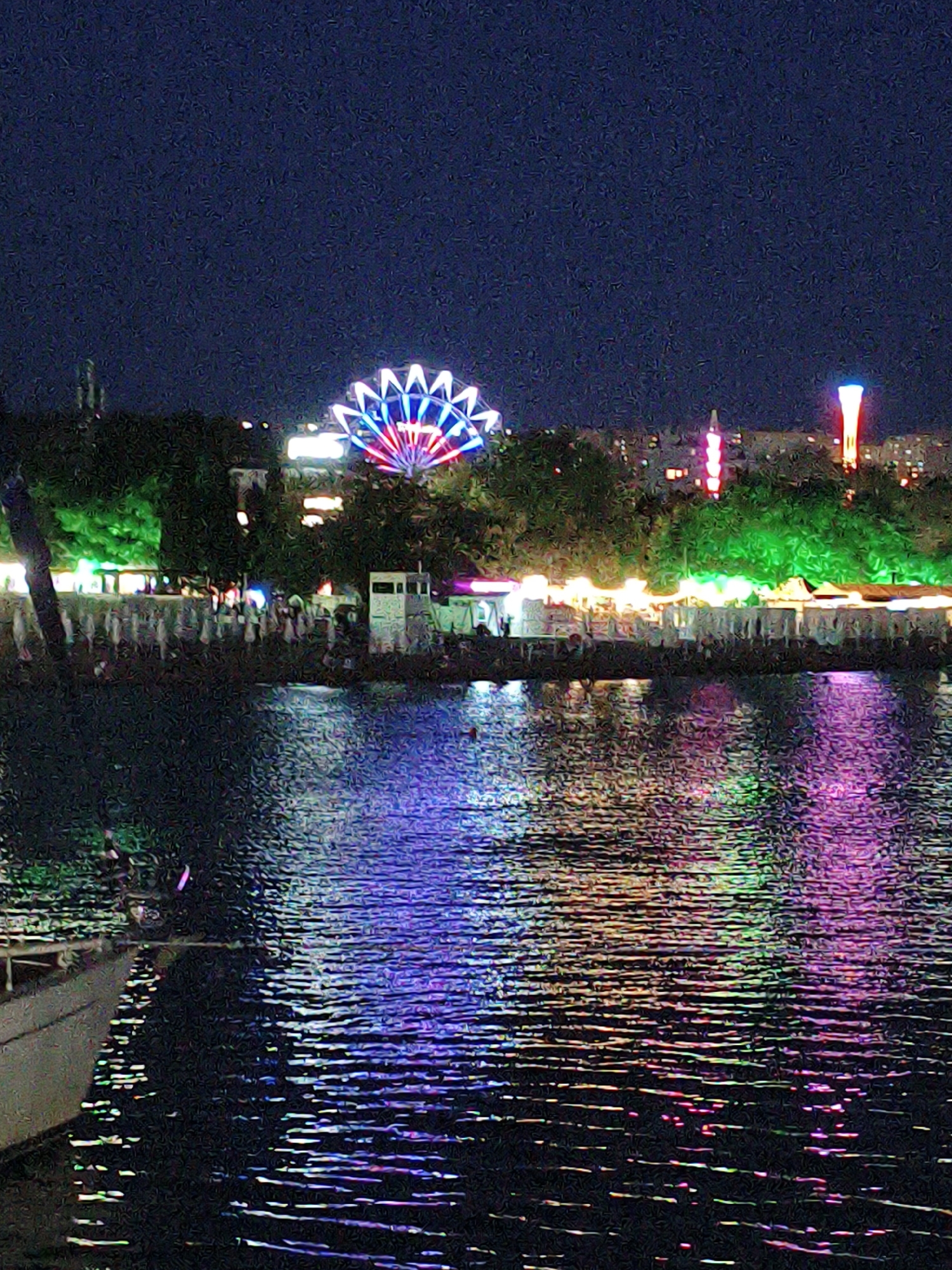 The image captures a nighttime scene at the Krinitsa, Russia waterfront. A Ferris wheel, illuminated with red, white, and blue lights, is visible in the background. Adjacent to the Ferris wheel are other illuminated structures and trees bathed in green light, suggesting a festive or amusement area. Several buildings are visible in the distance. The foreground is dominated by dark water, reflecting the colorful lights from the shore, creating a vibrant and shimmering effect. A portion of a boat is visible on the left side of the image. The overall atmosphere is one of nighttime entertainment and scenic beauty.
