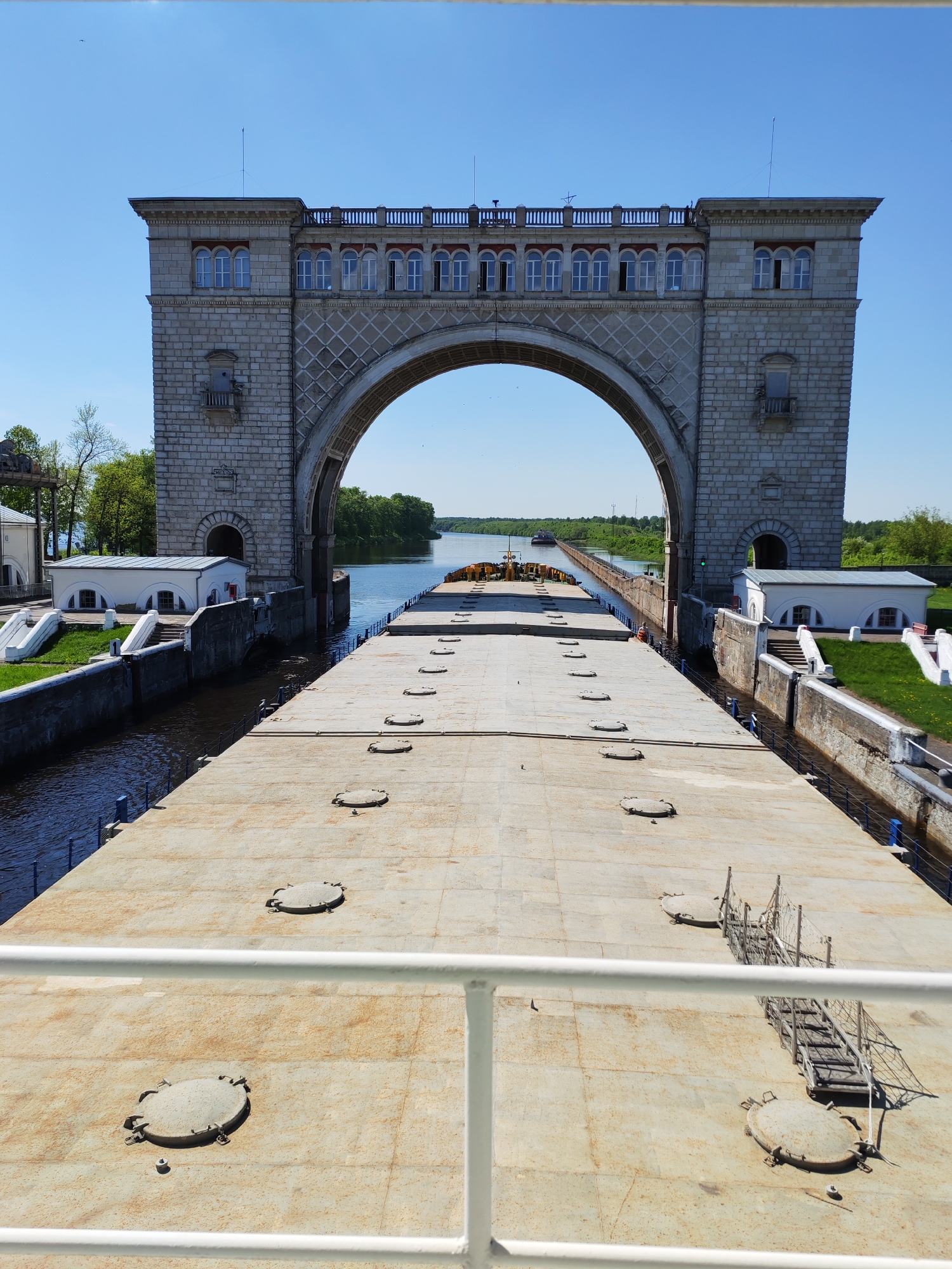 A full, eye-level shot depicts a massive, gray-stone arched structure framing a wide river waterway. The structure features multiple windows, decorative stonework, and prominent supporting pillars. A large, flat-topped barge occupies the foreground, its surface covered in numerous circular hatches. A white railing runs across the lower portion of the frame, suggesting the viewer is positioned on a ship or elevated platform.
On either side of the barge are parallel canal walls that appear to be part of a lock system. Small, white, one-story buildings sit adjacent to these walls, framed by manicured green grass. The river extends into the distance, flanked by lush green trees on either side. The sky is a clear, vibrant blue. The location is Unknown, Unknown.