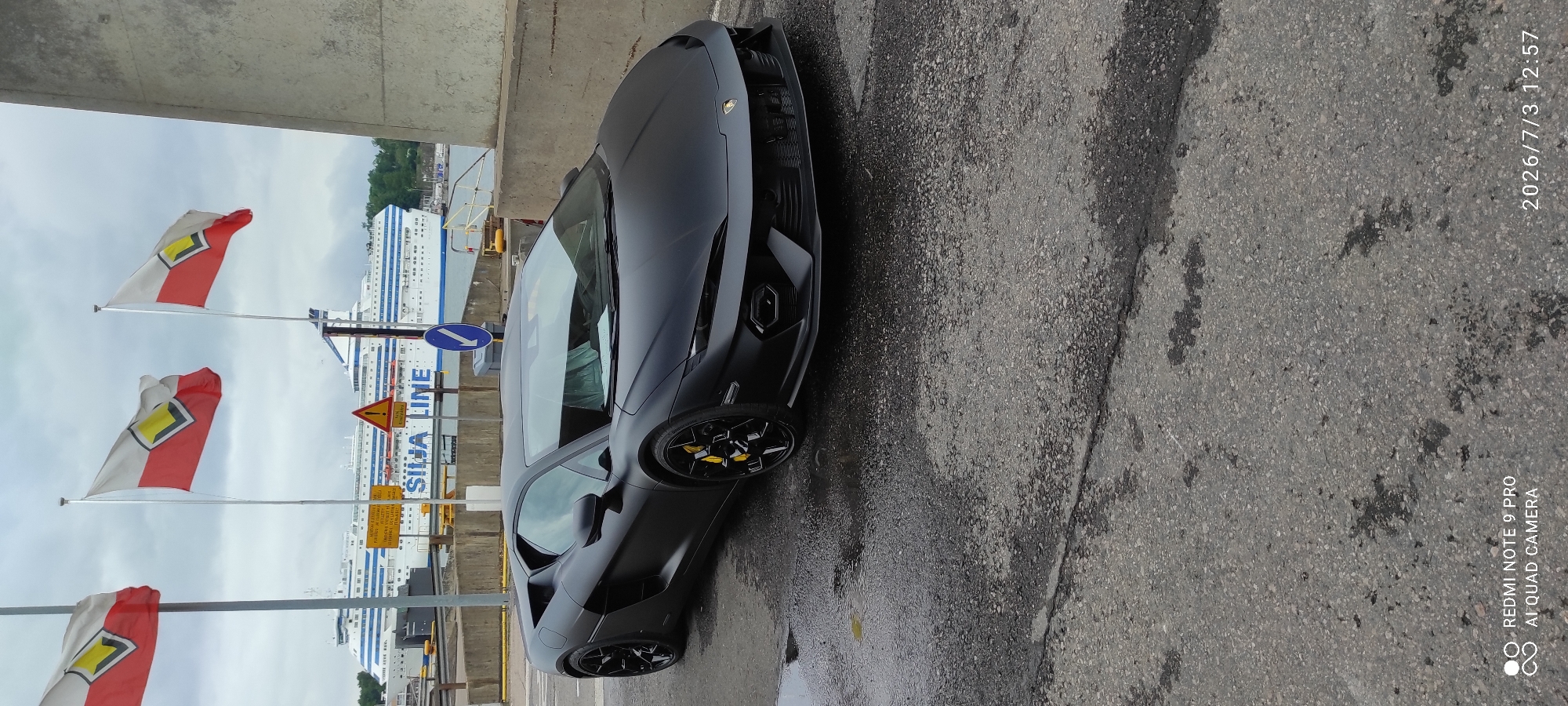 A matte black Lamborghini Huracan Performante is parked on a wet asphalt surface. The supercar is positioned in the foreground, facing slightly to the right of the camera, with its front bumper visible. Yellow brake calipers are visible behind the black multi-spoke wheels. In the background, a large white ferry with blue accents is docked. Three flags with a red and white pattern and a yellow diamond shape in the center are flying from poles to the left of the ferry. A concrete wall runs along the right side of the image. The sky is overcast. The lower right corner of the image contains a timestamp "2026/7/3 12:57" and device information "REDMI NOTE 9 PRO AI QUAD CAMERA". The location appears to be a port or waterfront area in Helsinki, Finland, suggested by the presence of the Silja Line ferry.
