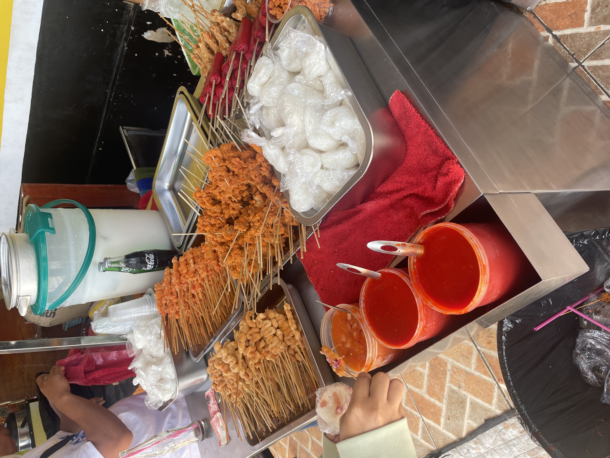The image depicts a food stall, likely in Davao City, Philippines, showcasing an assortment of deep-fried food items on skewers. In the foreground, two large plastic containers hold bright red dipping sauces, with spoons inserted. To the left, a person's hand is visible, holding a small white packet, possibly rice, and reaching towards one of the sauces. Behind the sauces, a metal tray is filled with numerous white packets, which also appear to contain food items. Further back, several metal trays are laden with skewers of food: one tray has numerous skewers of fried shrimp, another has skewers of what appears to be fried chicken or calamari, and a third tray contains skewers of red sausages. A large clear dispenser with a green lid, filled with a white liquid, is visible in the background, along with a Coca-Cola bottle and plastic cups. A black trash bin with pink straws is situated at the bottom left of the image. The stall appears to be outdoors, with tiled flooring visible in the foreground and a black wall with a yellow stripe behind the food display. The overall impression is of a bustling street food vendor.
