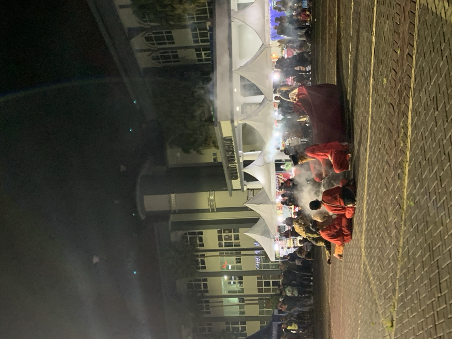 The image was taken at night in front of a large building in the city of Kecamatan Batu, Indonesia. The building has several windows and a sign that reads "Graha Pancasila". Several white tents are set up in front of the building. There are many people in the area and some are standing around the tents. In the center of the image, four people are sitting on the ground in front of a large red blanket. There is smoke around them. One person is wearing a black cap and a red shirt, while the other three are all wearing red shirts. The people are sitting in a circle. The ground is paved with gray bricks. There is a large parking lot and many people in the distance. The image was taken on a clear night with no clouds in the sky. The area is lit by streetlights.
