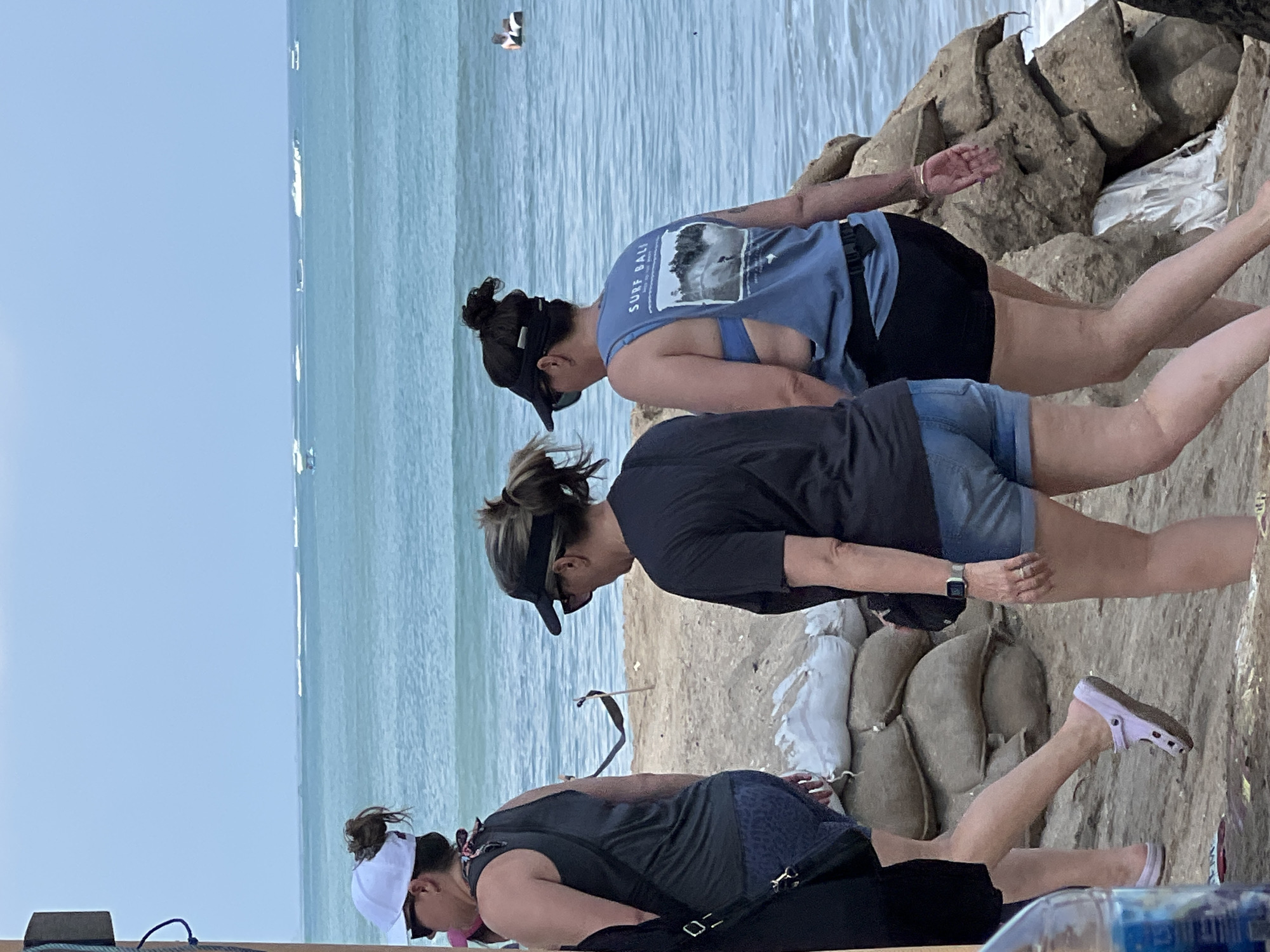 Three women walk along a sandy beach in Kuta, Indonesia. The woman on the left wears a white baseball cap, sunglasses, a dark tank top, and blue printed shorts. The woman in the middle wears a black visor, sunglasses, a black t-shirt, and denim shorts. The woman on the right wears a black visor, sunglasses, a blue tank top that reads "SURF BALI," and black shorts. They are walking towards the ocean, where several boats are visible on the horizon. Sandbags are piled on the beach in the foreground.