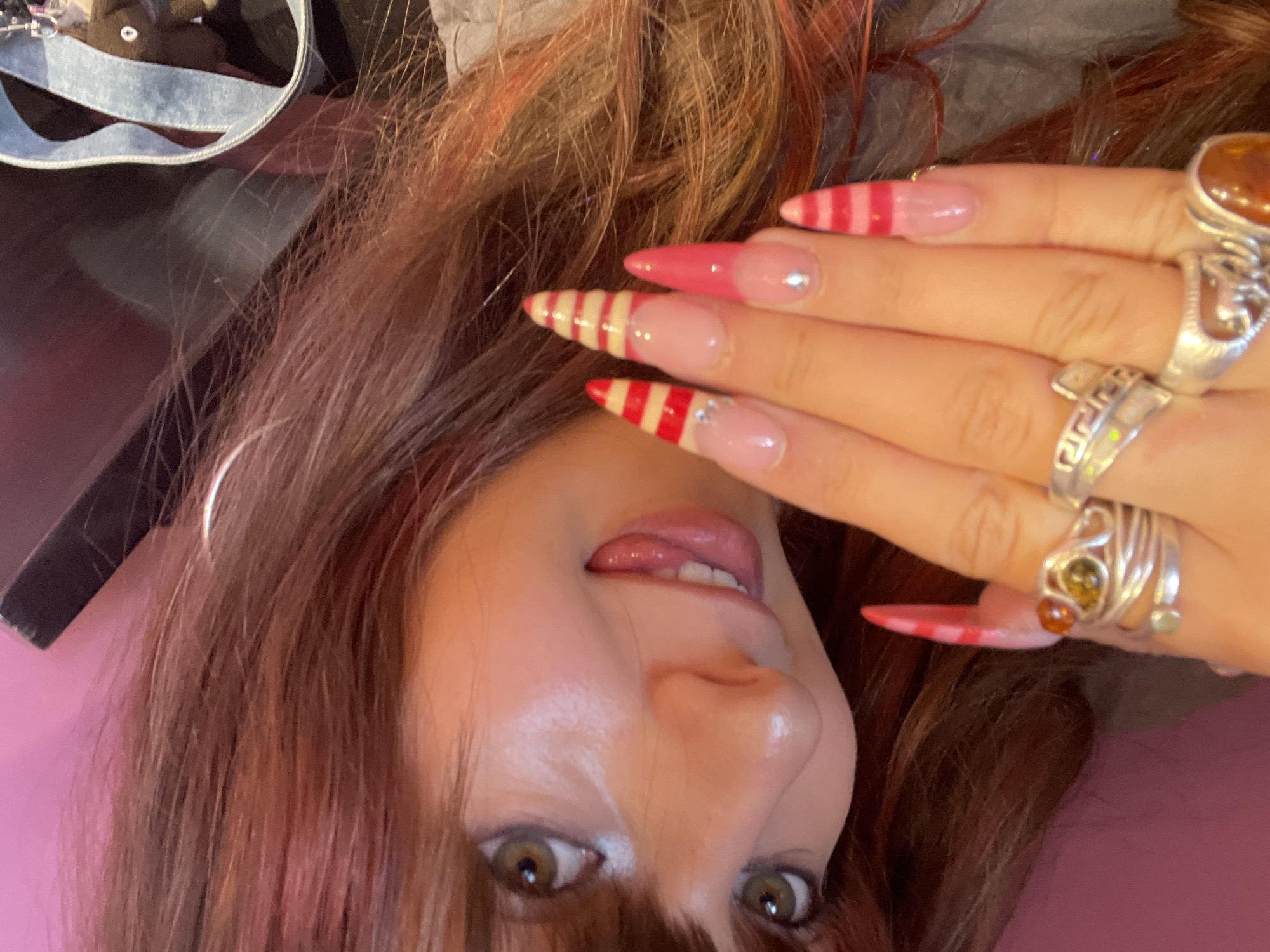 A young woman with reddish-brown hair and green eyes is shown in a close-up, selfies-style shot. She is looking towards the camera with her tongue sticking out slightly. Her nails are long and feature a pink and red striped pattern, with some decorated with small rhinestones. She is wearing several rings on her fingers, some of which appear to be silver with gemstones. The background is a solid pink color, with a portion of what appears to be a blue strap and a small brown stuffed toy visible in the upper right corner. The lighting suggests an indoor environment. The image is oriented sideways, with the woman's head on the left. The scene is devoid of any specific landmarks or indicators that would place it within the city of Raanana, Israel.