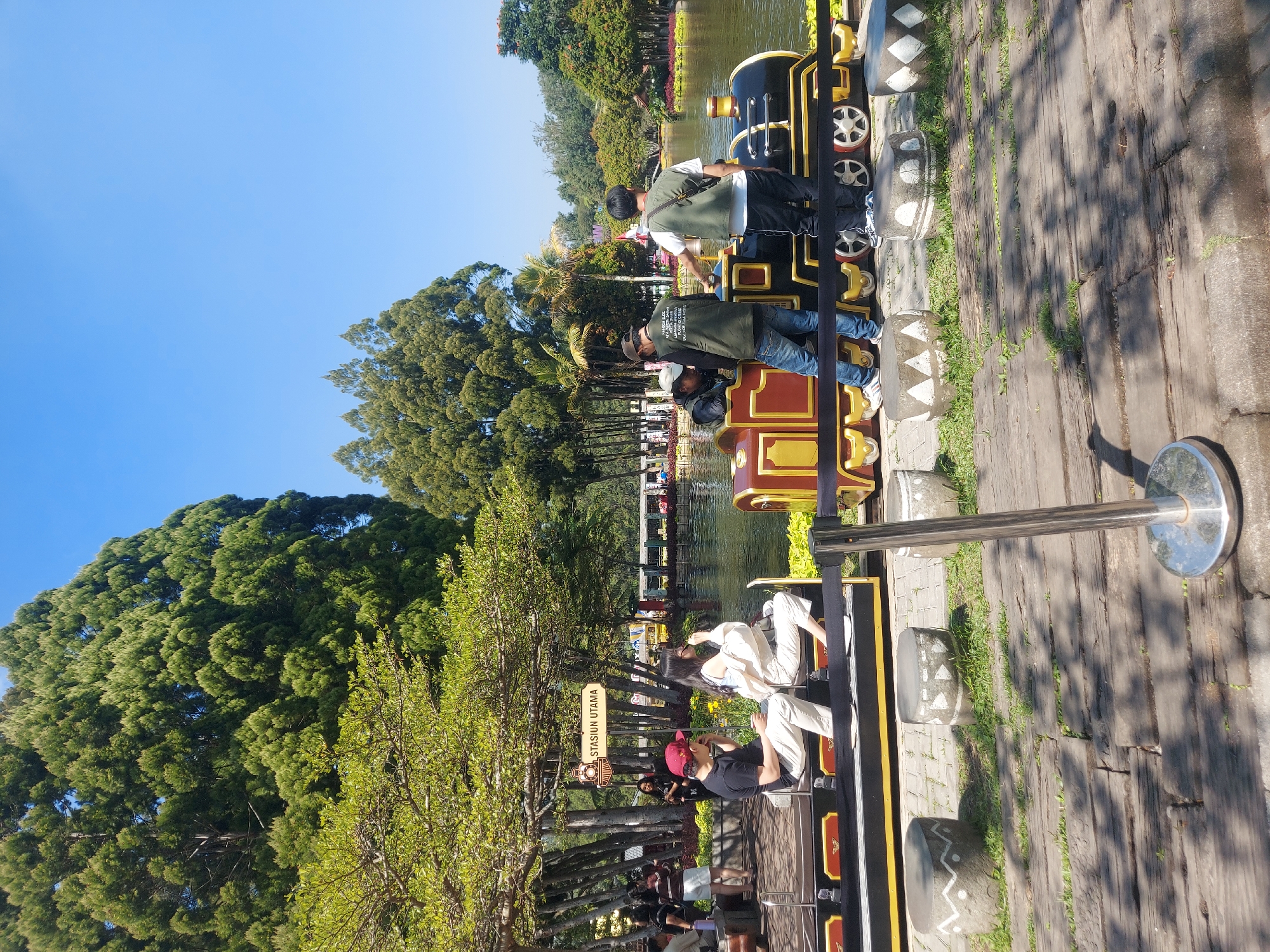 In Lembang, Indonesia, a miniature train attraction is set up next to a lake. The train, painted in red, gold, and black, is stationary, with several people interacting with it. Two individuals, a man in a black shirt and red cap and a woman in a white outfit, are seated on the train, facing forward. Behind them, three men are standing near the train. One man, wearing a dark jacket and jeans, is leaning towards the train. Another man in a green jacket and jeans is also near the train. A third man, in a white vest over a green shirt, is facing away from the camera, looking towards the lake. The background features a large body of water, lush green trees, and a clear blue sky. A sign above a group of trees reads "STASIUN UTAMA," meaning "Main Station." The ground is paved with wooden planks and cobblestones, with decorative stone stools placed along the path.