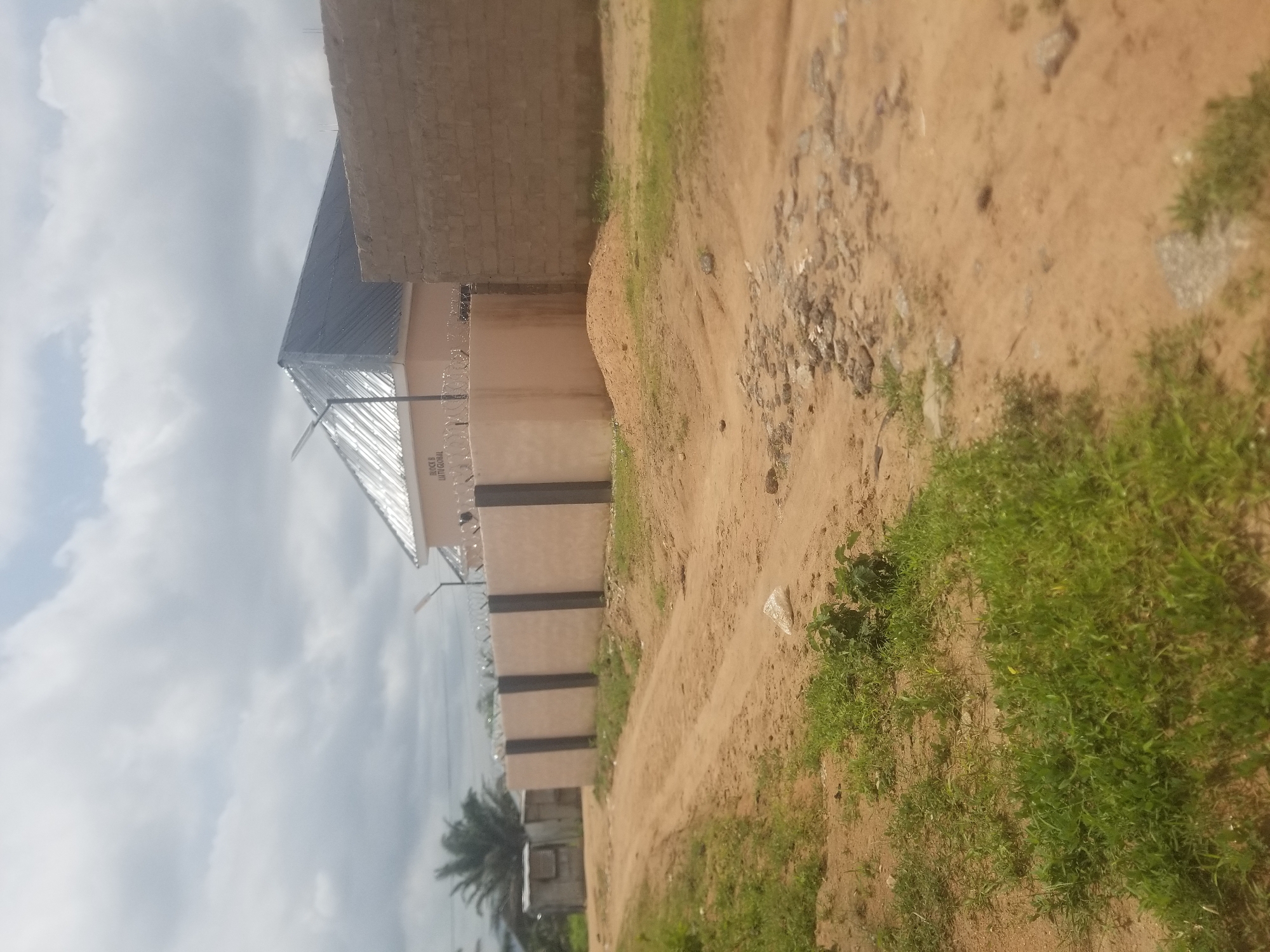 The image depicts a sunlit, outdoor scene in Jauro Yinu, Nigeria. A dirt road curves through the foreground, flanked by sparse green vegetation. In the middle ground, a beige concrete wall, topped with barbed wire, encloses a property. Beyond the wall, a building with a corrugated metal roof and a solar panel is visible. The building appears to be named "BLOCK B LIATU GLOBAL." To the right of this building, a wall constructed of tan bricks stands tall. The sky above is filled with white, fluffy clouds. A palm tree is visible in the far left of the image, and a small, gray metal shed is also present in the distance on the left side. The overall impression is of an undeveloped area undergoing construction.