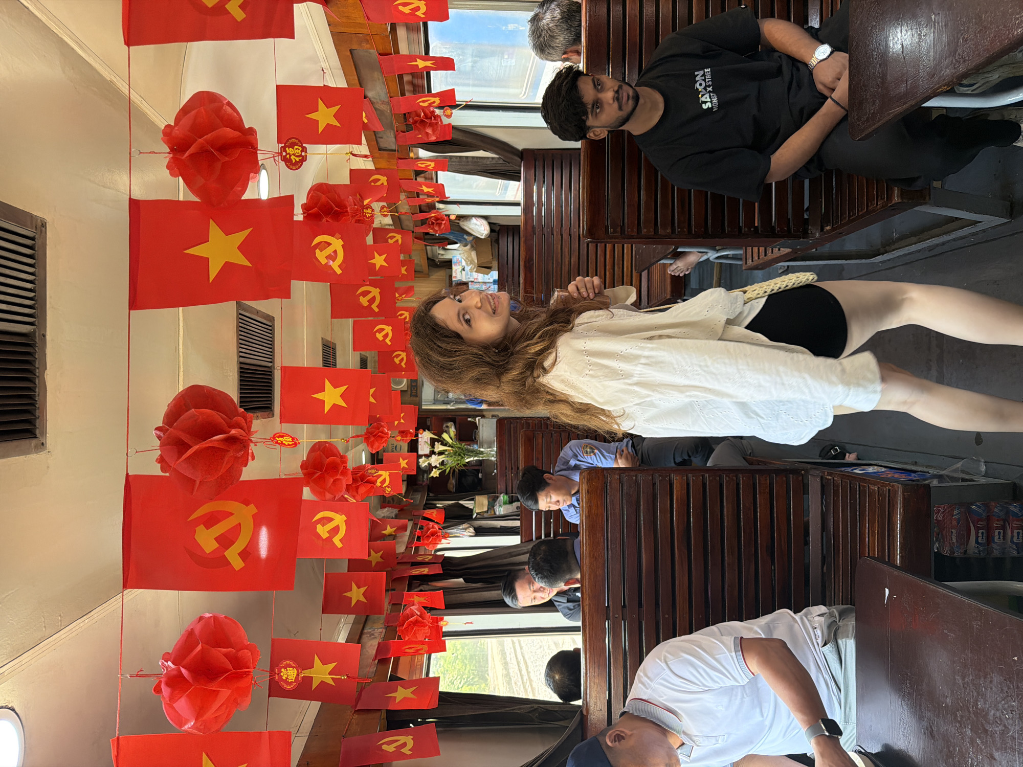 The photograph shows the interior of a train car in Huế, Vietnam. The car is adorned with strings of red Vietnamese flags and red paper lanterns. Several people are visible, seated in the car. A woman with long brown hair, wearing a white blouse, is smiling at the camera and holding a fan. A man wearing a black t-shirt sits across from her. Other people are seated further down the train car. The interior of the car is wood-paneled with wooden seating.