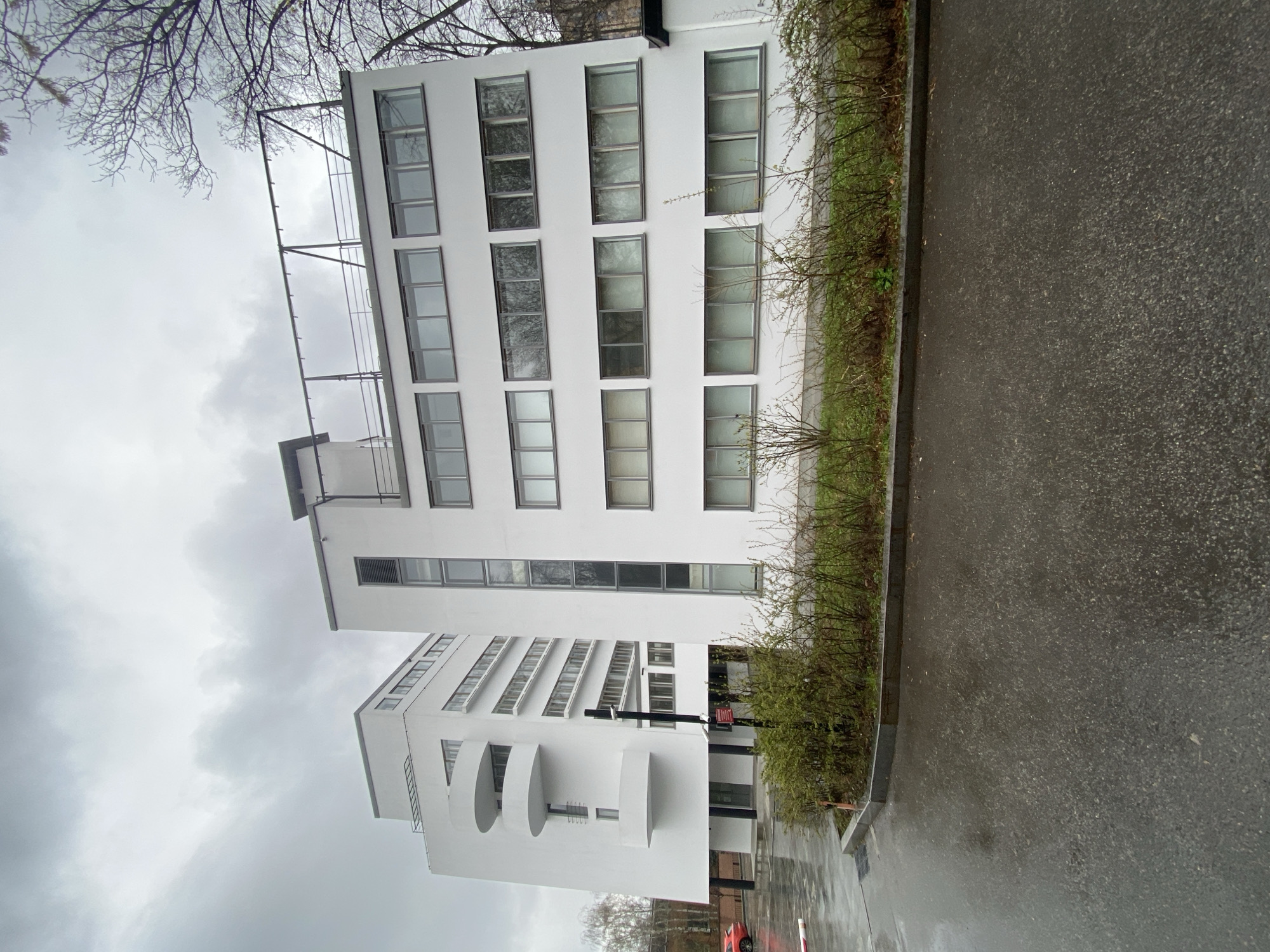 The image shows a white, modernist building with multiple rows of windows. The building appears to be located near a street with a curb and a row of bushes. The weather is overcast, and the pavement is wet, suggesting it may have recently rained. The top of the building has a skeletal frame structure.
