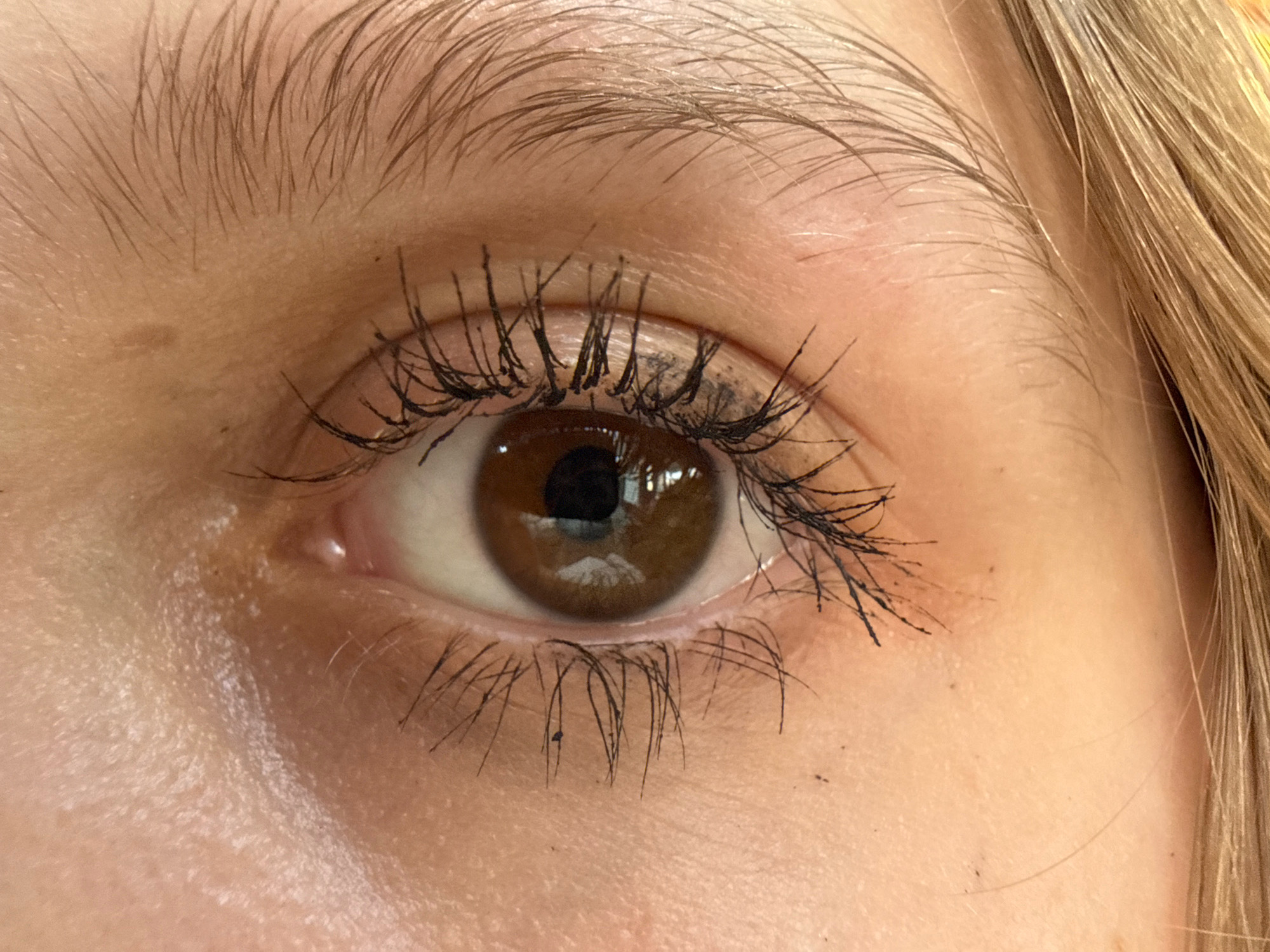 A close-up, eye-level shot focuses on a human eye with blonde hair visible on the right side of the frame. The eye is brown and appears to have black mascara applied to the eyelashes, with some smudging present. The skin around the eye is light-toned, and there are a few small dark spots on the skin, possibly blemishes or flecks of mascara. The lighting is bright, creating a slightly washed-out appearance. The image is an indoor, studio-like setting, as indicated by the consistent lighting and absence of any background elements that would suggest an outdoor or city environment. The exact location within a city is not determinable from the provided visual information.