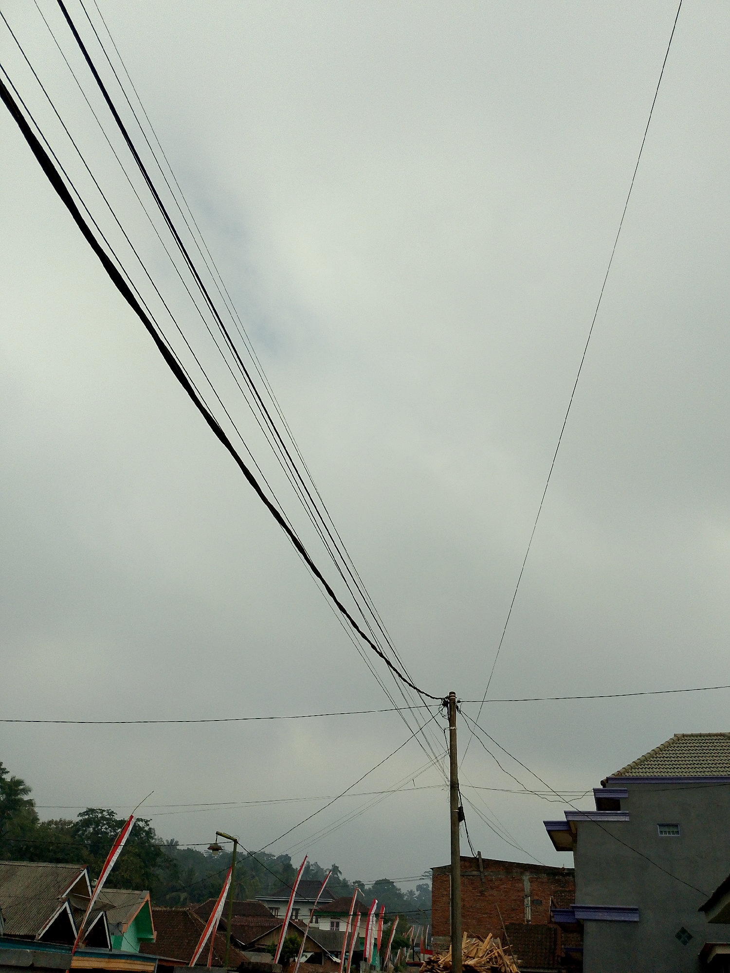The image depicts a cloudy sky over a residential area in Babadan, Indonesia. Multiple black power lines crisscross the frame, with a prominent cluster of lines originating from a wooden pole on the right side of the image and extending diagonally upwards to the left. In the lower part of the image, several houses with red and white Indonesian flags are visible. Some houses have tiled roofs, and one modern grey house with purple accents is situated on the right. Trees and foliage are present in the background. The scene appears to be a street view, with no people visible.