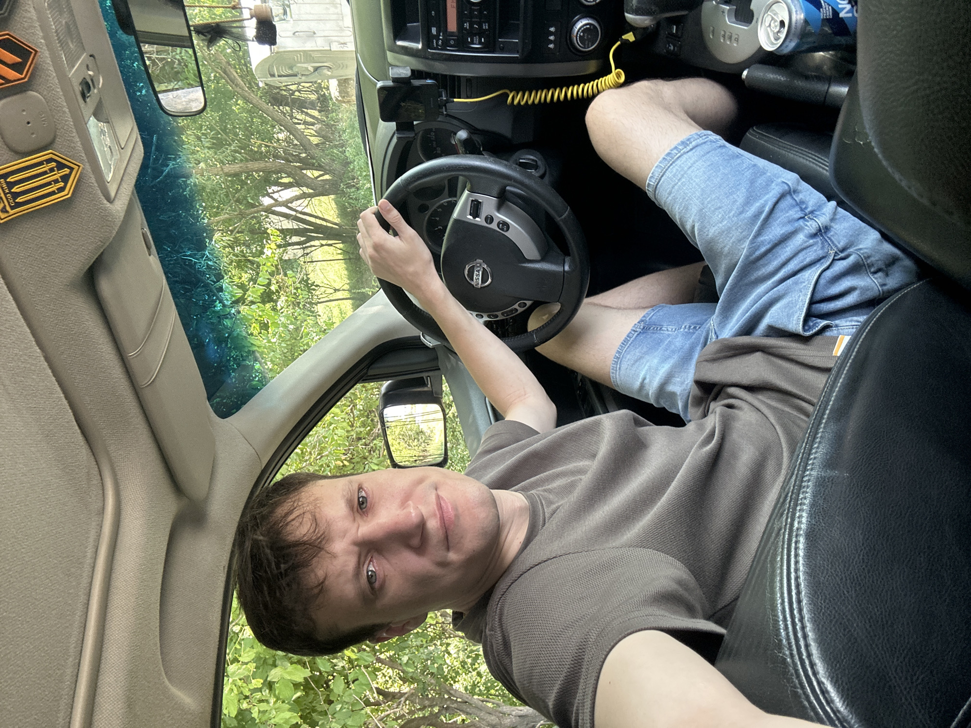 A young man is sitting in the driver's seat of a Nissan SUV. He is wearing a brown t-shirt and denim shorts. The man has short brown hair and light blue eyes. He is looking at the camera with a neutral expression. In the background, there is greenery, suggesting the car is parked outdoors. The interior of the car is visible, including the steering wheel, dashboard, and center console. A smartphone is mounted on the dashboard. A can of beverage is on the center console. The image appears to be taken in Makiv, Ukraine, given the mention of that location.