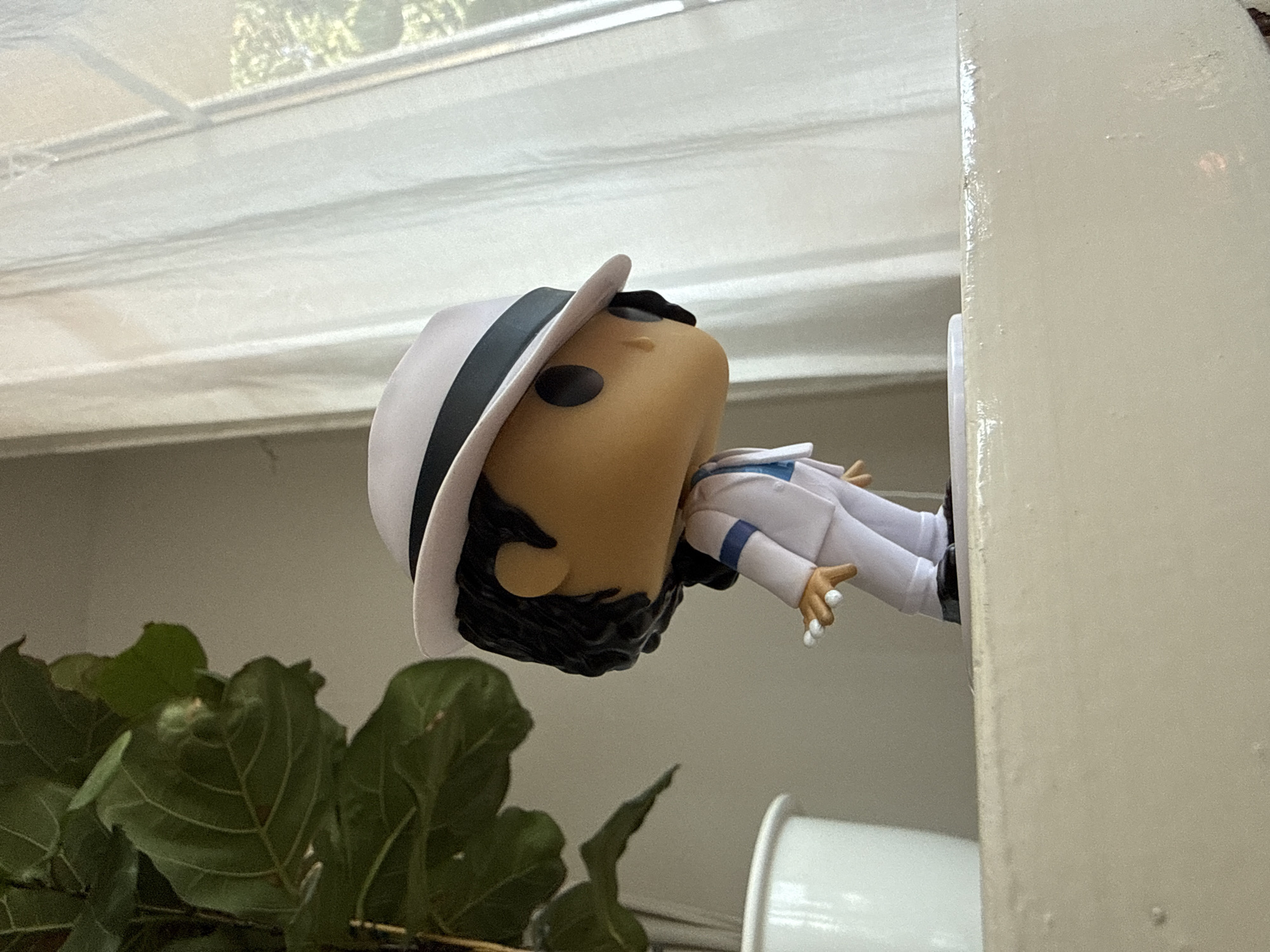 A Michael Jackson Funko Pop figure, dressed in a white suit with a white hat, stands on a light-colored surface. A portion of a large green leafy plant in a white pot is visible to the left of the figure. To the right of the figure, white sheer curtains hang near a window. The background is a neutral, light-colored wall. The image is taken from a low angle, looking up at the figure. The setting is indoors, likely in a domestic environment. There is no indication of the city of Bad Sachsa, Germany.