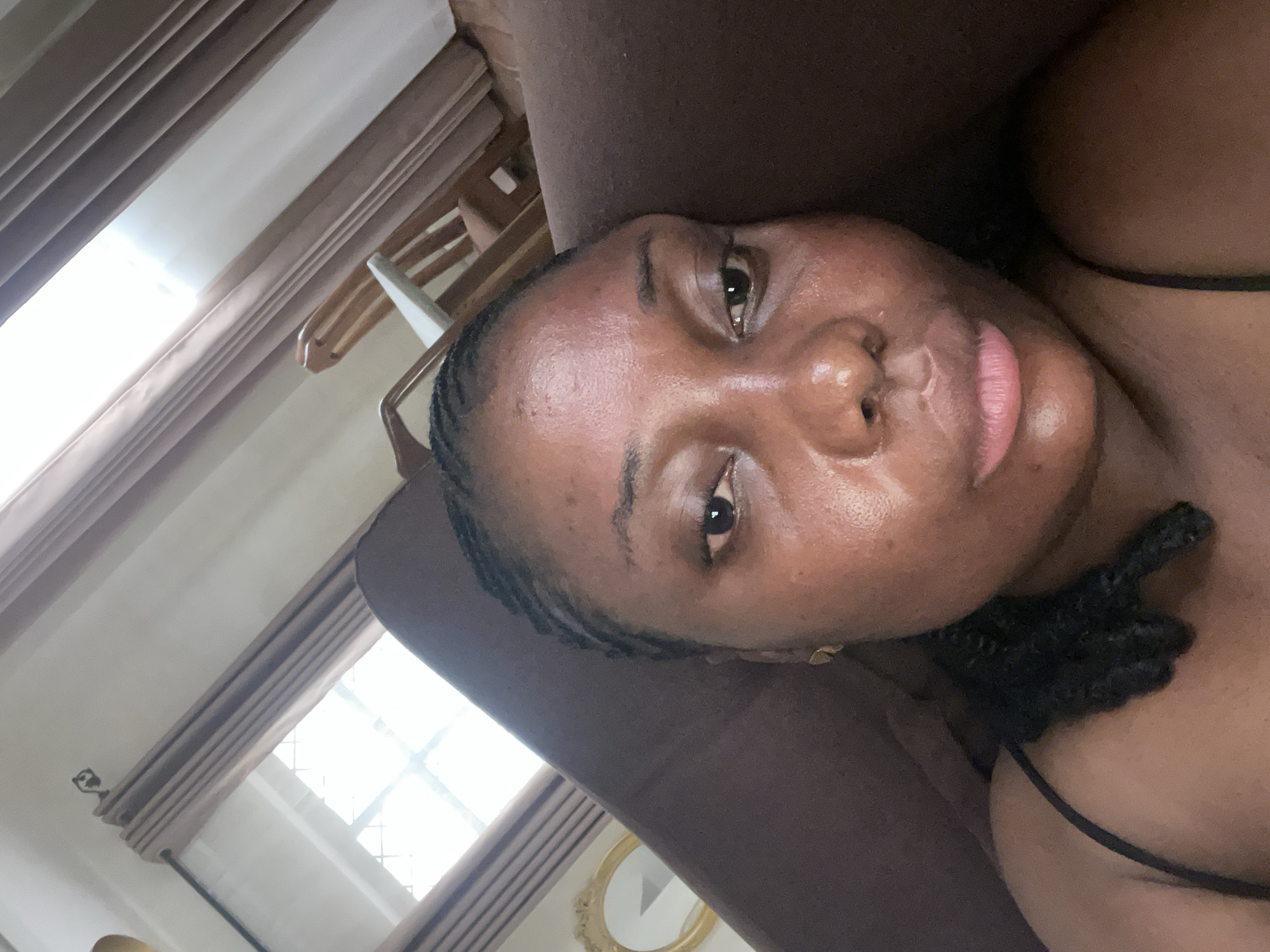 The rotated image presents a close-up portrait of a woman with dark skin, likely taken in a selfie style while she is lying down. Her head is tilted, and she gazes directly at the viewer with a neutral expression. Small, dark braids frame her face, and her skin has a slight natural sheen, revealing some texture around her forehead and cheeks. Her lips are a light pinkish-brown. She is wearing a dark top with a thin strap visible on her shoulder.
She appears to be resting on a soft, dark brown surface, possibly a pillow or couch. In the background, to what would be her upper left if the image were upright, a bright window with a grid pattern is visible. Abundant natural daylight streams through, suggesting it is daytime. Light-colored, possibly sheer, curtains are drawn back from the window. Behind the curtains, subtle outlines of wooden furniture, resembling coat hangers or a chair, can be discerned. To her lower left, a circular, gold-framed ornate mirror or decorative item is partially visible on the wall.
The setting is an indoor residential space, potentially a bedroom or living room in Jos, Nigeria. There is no visible text in the image.