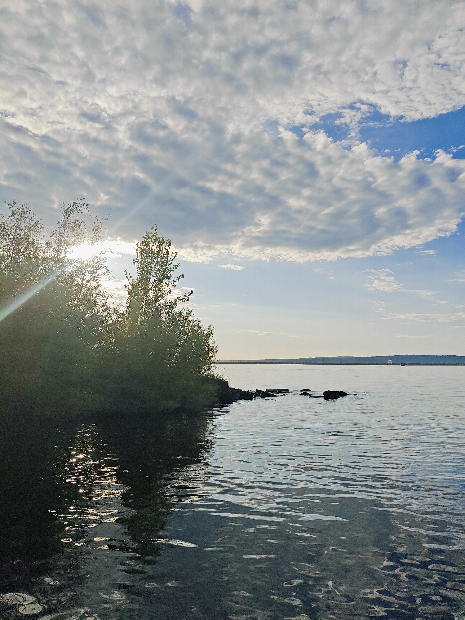 A wide shot captures a serene natural landscape under a partially cloudy sky. The sun shines brightly, partially obscured by the leaves of lush green trees on the left side of the frame, casting sunbeams and dappled light onto the water. The water itself is calm, reflecting the sky and the foliage. A small cluster of dark rocks juts out of the water near the shore. In the distance, a flat horizon line separates the water from a distant landmass, with a faint structure visible on the horizon. The scene appears to be a body of water with a vegetated shoreline, possibly a lake or river. The location is Chaikovsky, Russia.