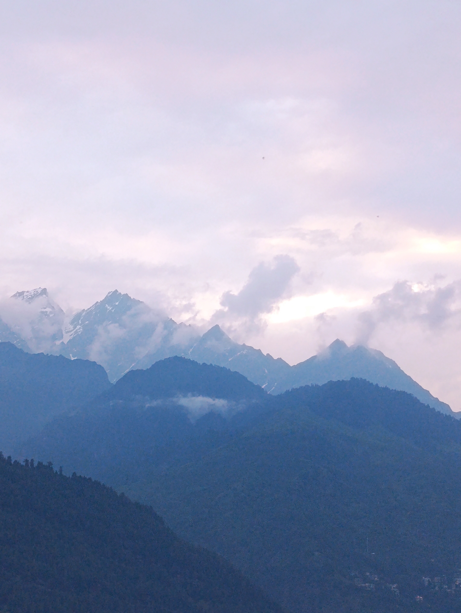 A panoramic view of majestic mountains is captured, with layers of peaks fading into the distance under a soft, overcast sky. The foreground is dominated by the lush green slopes of a mountain, densely covered in trees. Further back, a series of rugged mountain ranges rise, their peaks partially obscured by wispy clouds and mist. Some of the higher peaks are snow-capped, glistening faintly in the diffused light. The sky above is a pale canvas of pinks and grays, with hints of brighter light peeking through the clouds, suggesting either sunrise or sunset. The overall atmosphere is serene and tranquil, highlighting the grandeur of the natural landscape. This scene is likely located in or near Vashisht, India, given its mountainous terrain.