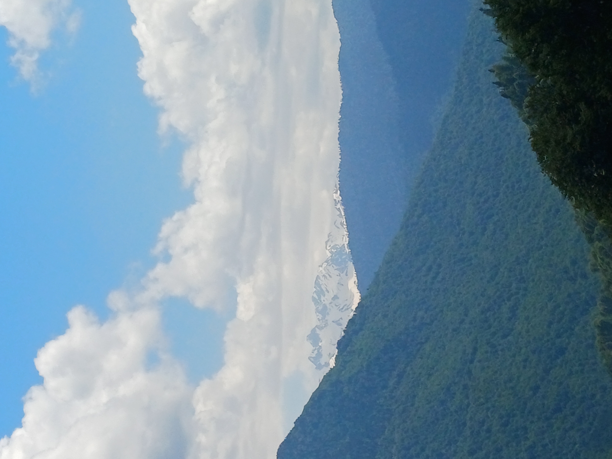 The image depicts a vast mountain landscape under a partly cloudy sky. The foreground is dominated by the lush green slopes of a mountain covered in dense forest. The middle ground features a range of mountains in a deep blue hue, with distinct layers of peaks receding into the distance. In the background, a majestic mountain range with snow-capped peaks stands out against the lighter blue sky. The sky itself is a vibrant blue, dotted with fluffy white clouds. The scene appears to be a serene, natural environment, likely part of the mountainous terrain surrounding the city of Estosadok, Russia, known for its alpine scenery. There are no people or identifiable man-made objects in the image.