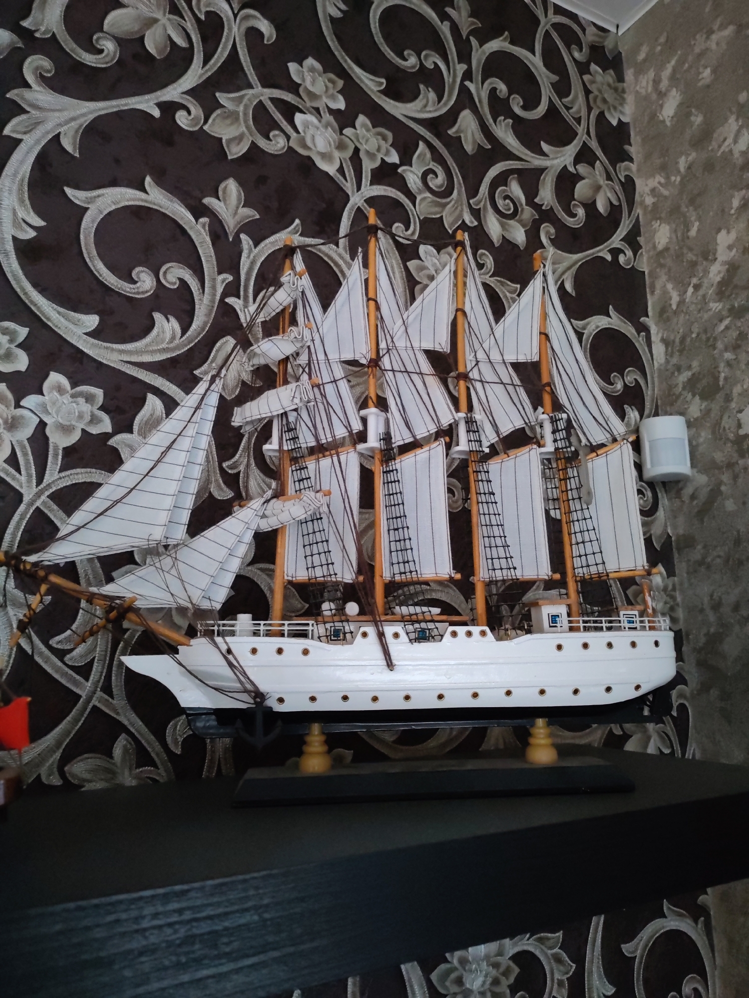 A close-up, eye-level, medium shot shows a detailed model ship on a black shelf. The ship is white with multiple sails, masts, and rigging. It is mounted on a black base with wooden stands. The background features dark brown wallpaper with a silver floral pattern and a textured grey wall on the right. A small white motion sensor is visible on the grey wall. The lighting is ambient, coming from the front and left of the scene. There are no people or animals present.