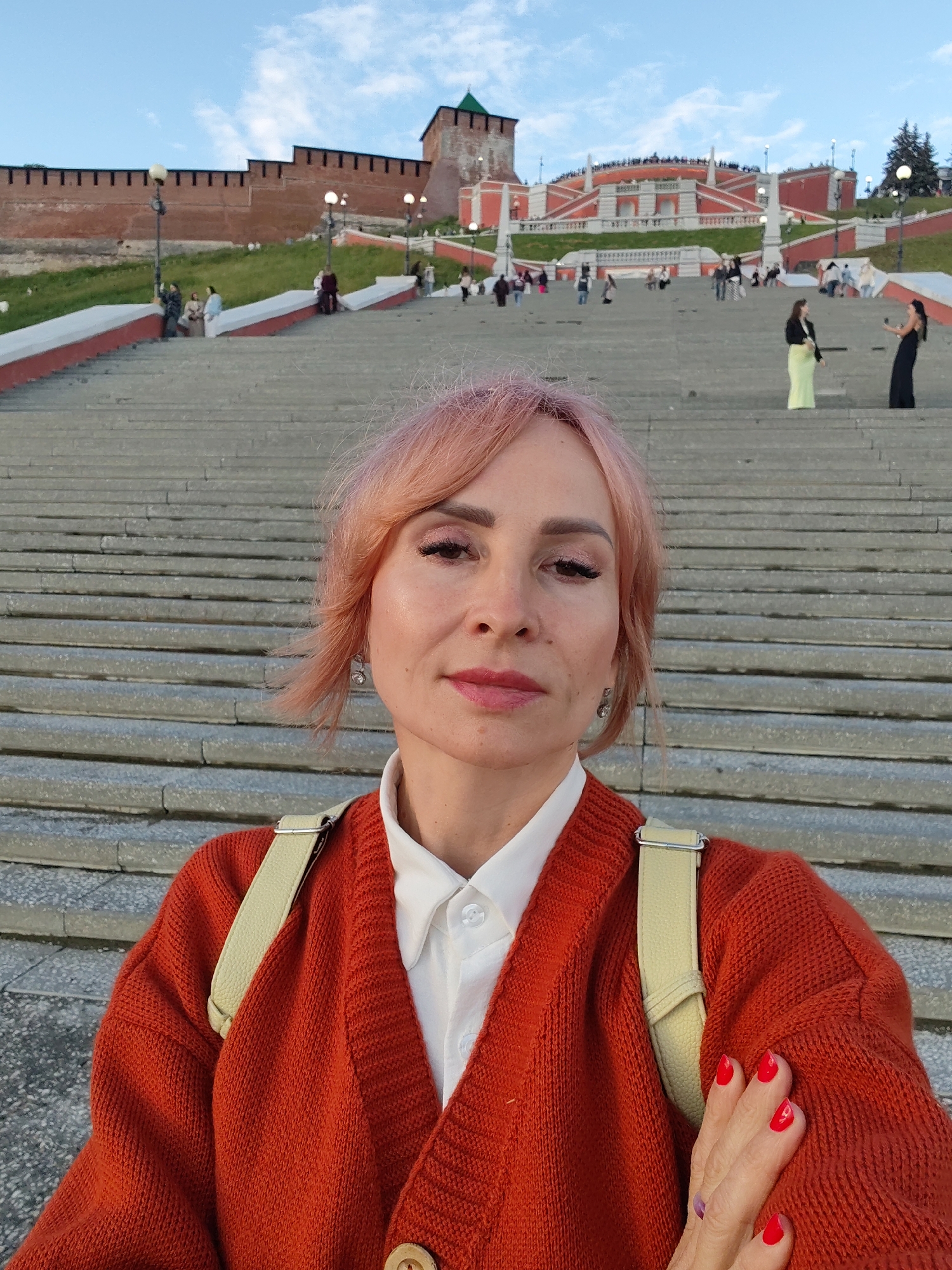 This is a medium shot selfie of a woman with pinkish-blonde hair, wearing a bright orange cardigan over a white collared shirt. Her left hand, adorned with bright red nail polish, is resting on her chest. She is positioned on a wide, outdoor stone staircase, with many steps receding into the background. In the mid-ground and background, a large group of people are visible ascending and descending the stairs and milling about. The setting appears to be a historical site with red brick walls and a tower visible on a grassy hill behind the staircase, suggesting a fortress or kremlin. The architecture of the buildings in the background, with red and white accents and a distinct tiered design, further supports this. The sky is a clear blue with a few wispy clouds, indicating it is daytime, likely late afternoon or early evening given the soft lighting. The overall mood is relaxed, with people enjoying the location. There is no visible text in the image.