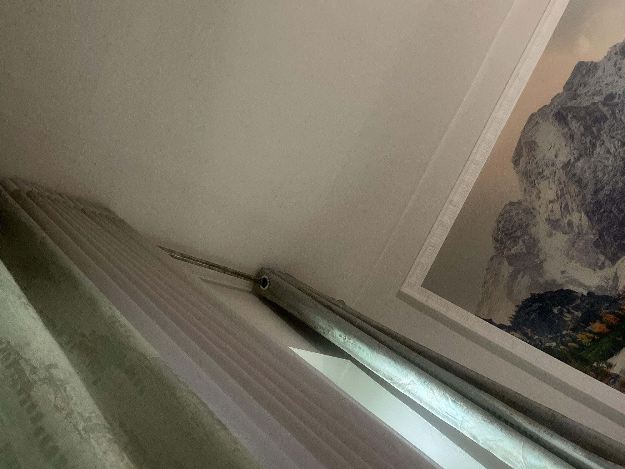 The image shows an interior view of a room. A light green patterned curtain hangs from a curtain rod, partially obscuring a white wall and a framed picture of a mountain landscape. The curtain rod is silver and has visible grommets. A sliver of light shines through a gap between the curtain and the wall. The ceiling is white with some faint markings. There is a decorative molding running along the top of the framed picture. The scene appears to be taken from a low angle, looking upwards. The image does not provide any information about the location in Bishkek City, Kyrgyzstan.
