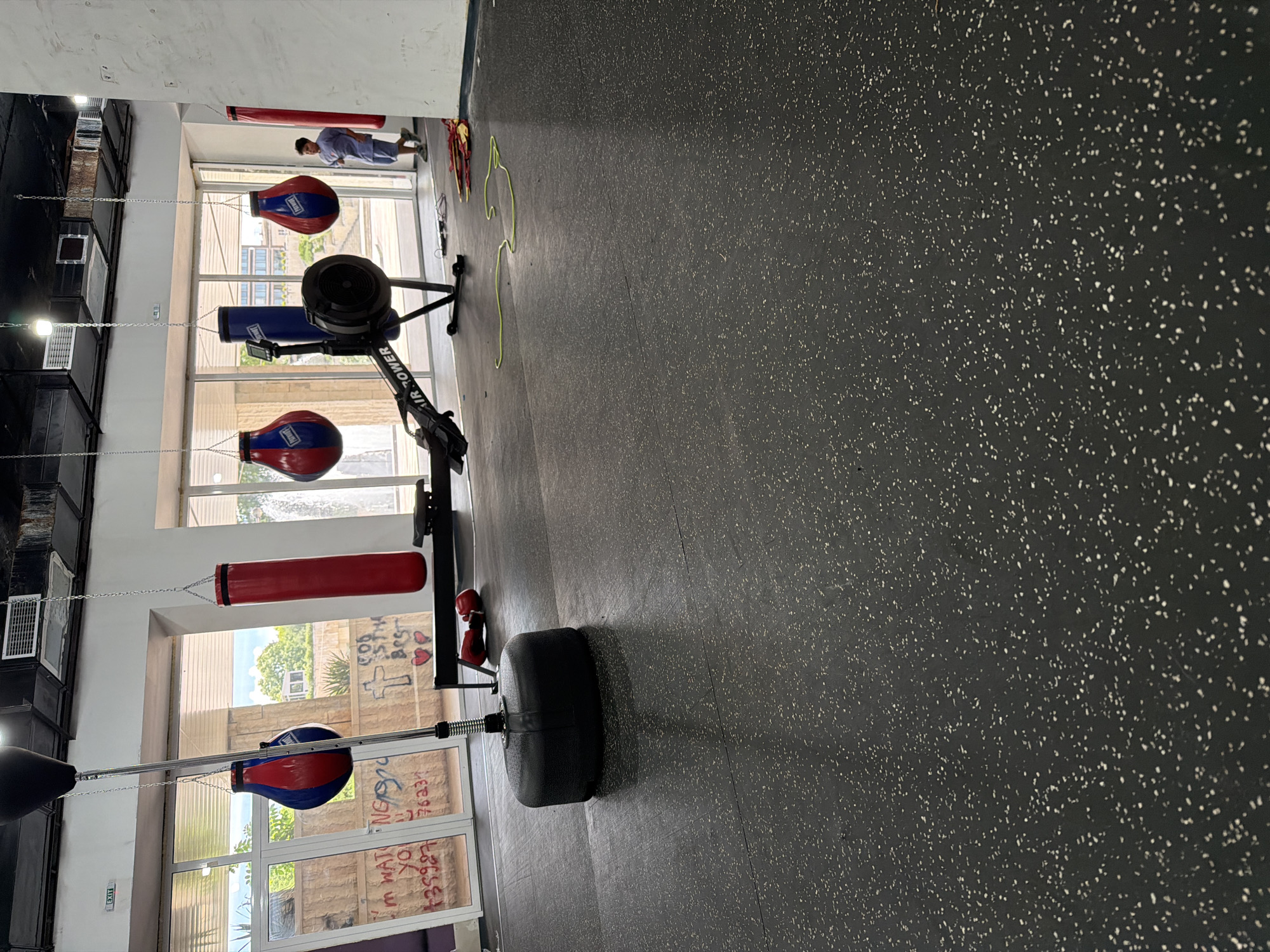 A gym with boxing equipment and a rowing machine is depicted in Blagoevgrad, Bulgaria. The gym features a black rubberized floor with white speckles. In the foreground, a black, rounded punching bag base is visible. To the left, a speed bag is suspended from a metal pole. Hanging from chains are two red and blue boxing bags, and a single red boxing bag. A black rowing machine with "AIR TOWER" written on it is positioned in the middle of the gym. A young man wearing a light blue t-shirt and shorts stands near the back right of the image, looking towards the rowing machine. Red boxing gloves lie on the floor near the rowing machine. A green resistance band is also present on the floor. The gym has large windows offering a view of trees and buildings outside. Graffiti, including a cross and text, is visible on a wall through the windows.