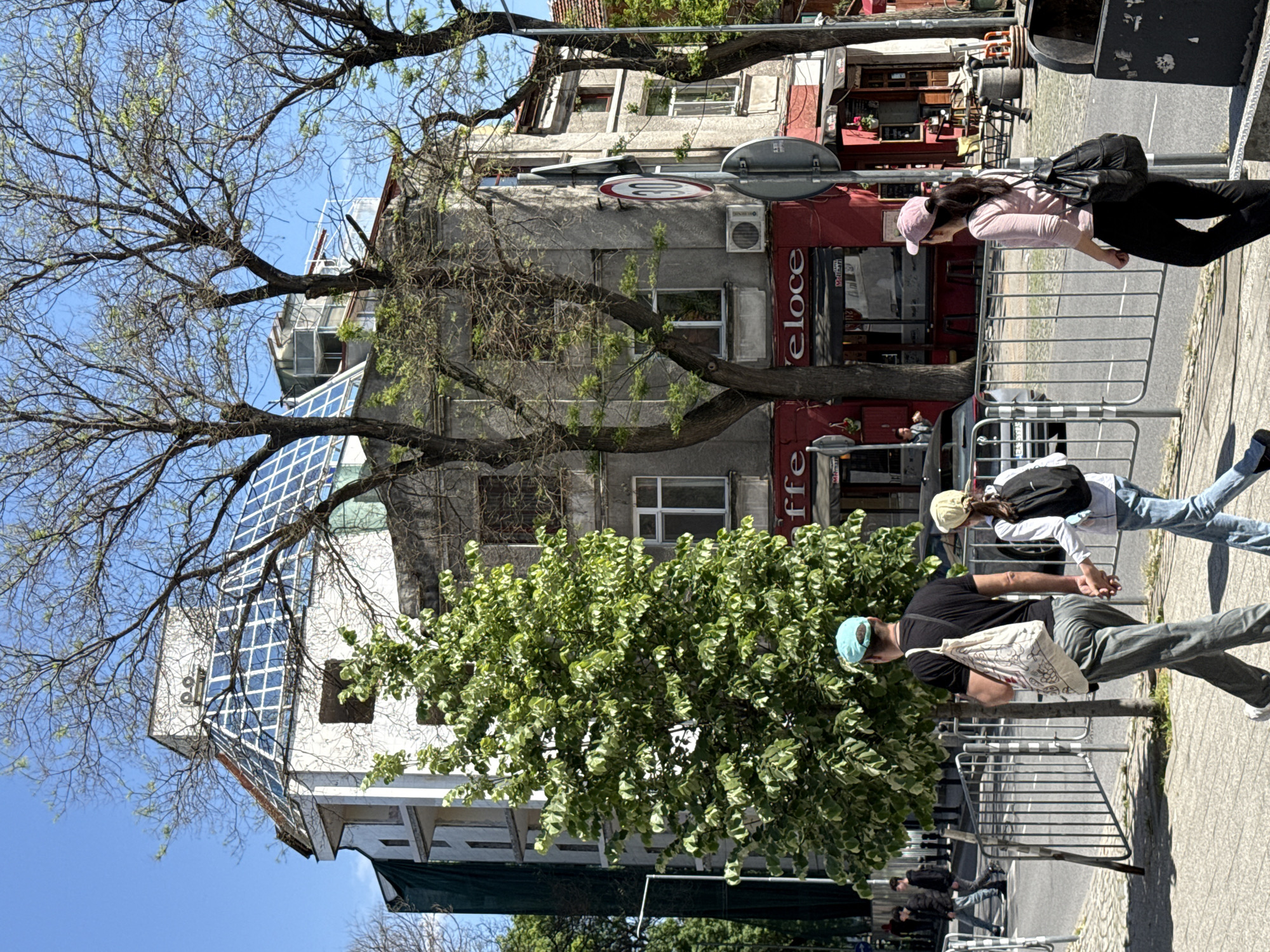 The image captures a street scene in an urban environment, showing a mix of architectural styles and pedestrian activity. In the foreground, a metal barrier runs parallel to the sidewalk. Two pedestrians, a male and female, are walking along the sidewalk behind the barrier. The man is wearing a black t-shirt and carrying a shoulder bag. The woman has a white shirt and a backpack.
Behind them is a building with a red-awninged storefront labeled "Caffe Veloce." Above that, the building has a gray facade with multiple windows and signs. To the left is another building featuring a more modern, glass-paneled design. Tall trees with partially budding leaves and green foliage frame the buildings, with a clear blue sky visible above.
The overall setting suggests a city center location with a blend of commercial and residential buildings and a mix of old and new architectural elements.