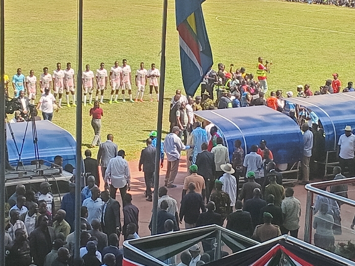 The image captures an outdoor scene at a sports stadium or football pitch, bathed in bright daylight under what appears to be clear skies.
In the mid-ground, a team of male football players, dressed in light-colored (likely white or very light pink) jerseys, shorts, and light-colored boots, are lined up in a row on the green grass. The word "KENYA" is visible on their jerseys, identifying them as representing Kenya. A woman in a red top and dark trousers walks in front of the lined-up players. To the left, a vehicle with speakers mounted on its roof has "KENYA" visible on its side.
Two blue-covered dugouts or shelters are prominent features across the mid-ground, with numerous people, including officials, security personnel, and spectators, gathered around and inside them. A man wearing a red vest and holding a camera or video recorder stands near one of the dugouts.
In the foreground, a large crowd of people, many dressed in smart casual or semi-formal attire, are gathered, suggesting a significant public event. A flag with blue, yellow, and red elements is flying from a pole at the top left, while a banner in the bottom right corner displays the colors of the Kenyan flag (black, red, and green).
The overall activity suggests a pre-match ceremony or an official visit during a football event, possibly involving dignitaries, in Siaya, Kenya, given the context and visible national symbols. The scene is well-attended and appears organized.