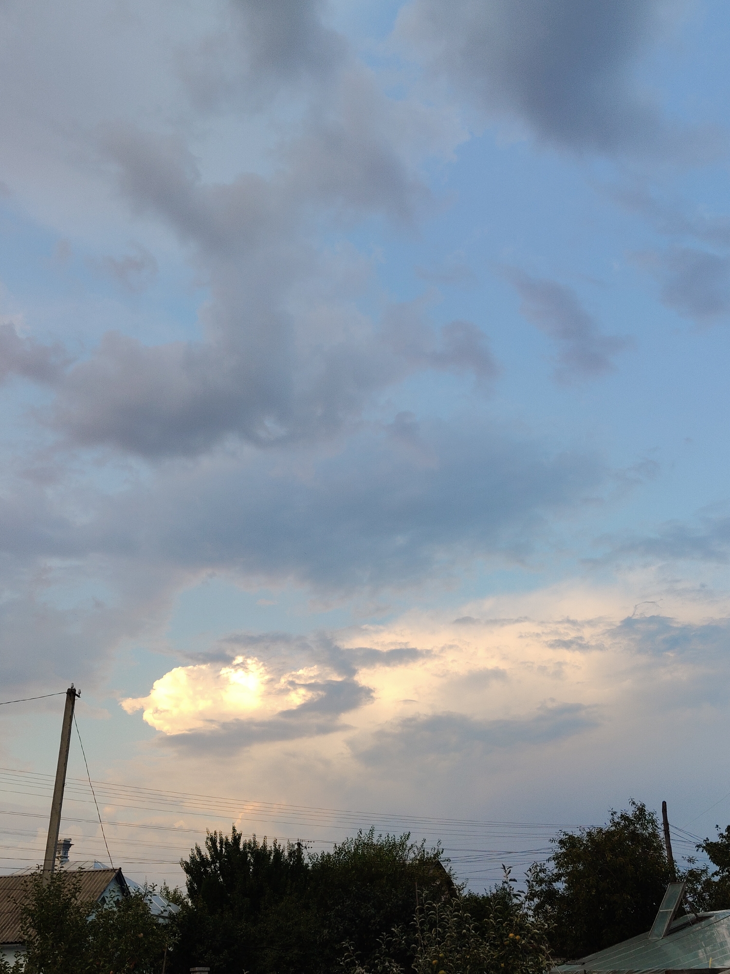 The image depicts a rural or suburban sky with scattered clouds. In the foreground, the rooftops of several houses and a dense line of trees are visible. A utility pole with electrical wires runs diagonally across the left side of the frame. The sky is a mixture of light blue and grey clouds, with a bright, illuminated cloud formation towards the center left, suggesting sunlight breaking through. The scene appears to be captured during daylight hours, likely in the late afternoon or early evening due to the quality of light. No people or specific actions are identifiable. The location is identified as Cherkasy, Ukraine.