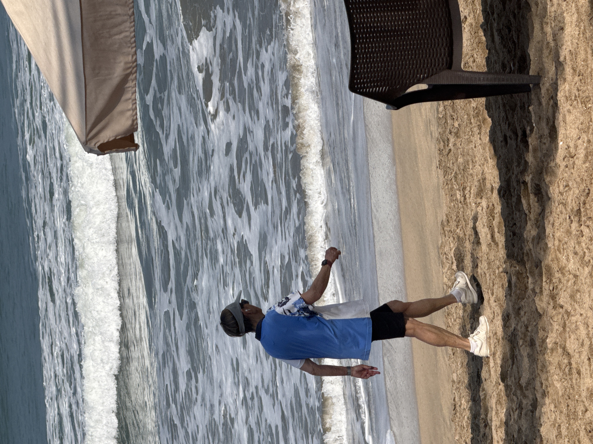 A person walks on a sandy beach in Kuta, Indonesia, with waves in the background. The individual, wearing a blue and white shirt, black shorts, white sneakers, and a visor, is captured from the rear, facing left. A brown plastic chair and the corner of a tan umbrella are visible in the foreground on the right. The ocean exhibits white foam and ripples under a bright sky.