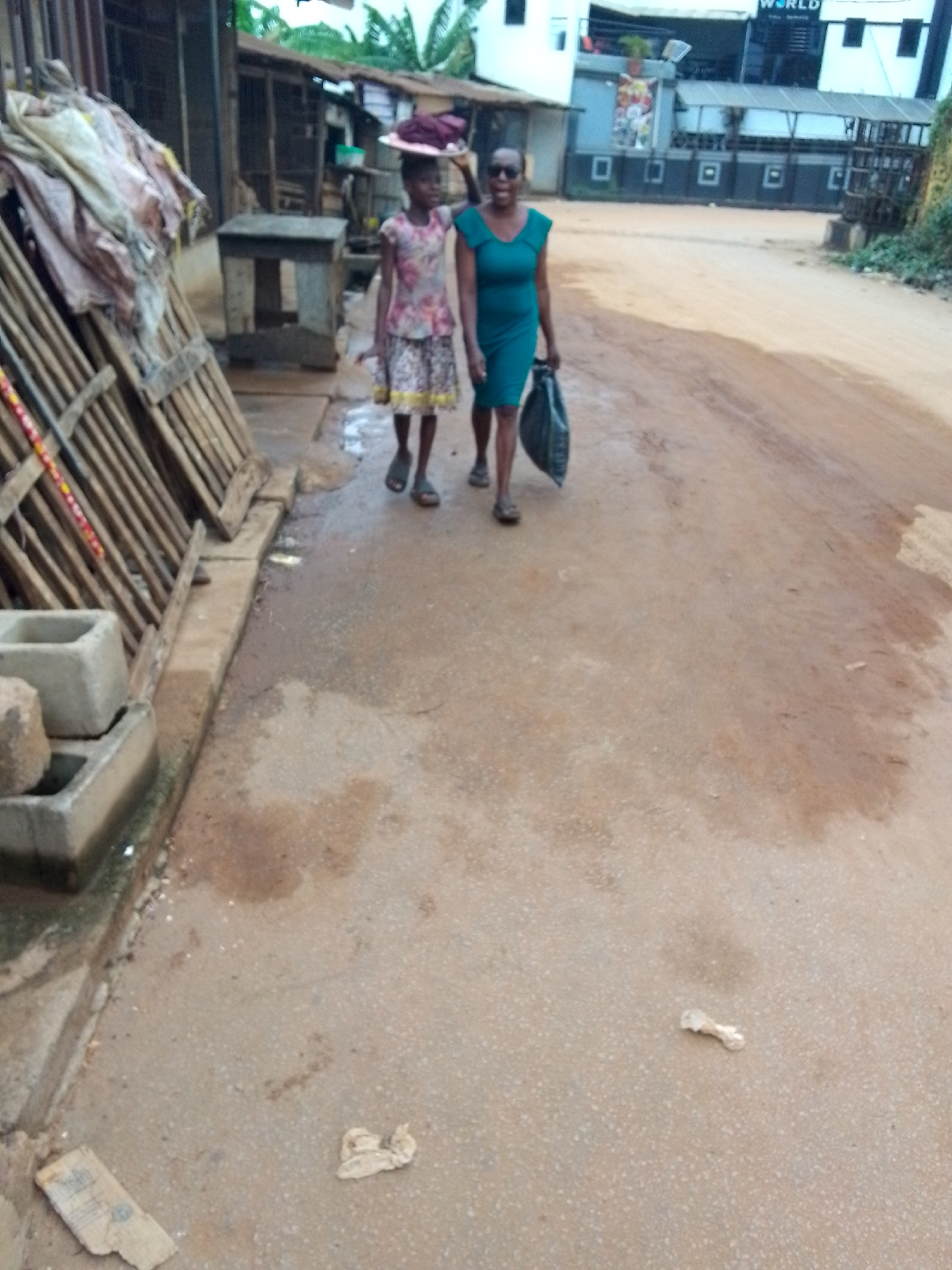 Two females, an adult and a child, walk along an unpaved, reddish-brown dirt road in what appears to be a semi-urban or urban area, identified as Onitsha, Nigeria. The adult, on the right side of the frame, wears a dark teal or green dress and sunglasses, carrying a black bag in her left hand. She is smiling and looking towards the camera. The child, walking slightly ahead and to the left of the adult, is dressed in a floral-patterned top and skirt, and carries a pink or purple hat or cloth on her head. She looks towards her right.
To the immediate left of the walking path, stacked wooden planks or crates are visible, alongside two grey concrete blocks resting on a damp, lighter-colored concrete strip. This strip runs parallel to the dirt road, which itself shows signs of dampness or recent rain with scattered wet patches. Small pieces of paper or plastic litter are visible on the ground.
In the background on the left, simple structures, possibly huts or stalls with corrugated iron roofs, are seen. Further back and to the right, a more modern, two-story white building with dark windows and accents is prominent. Part of a sign on this building reads "WORLD". Green foliage and trees are visible beyond the structures. The scene suggests daytime, possibly overcast, with an active street environment.