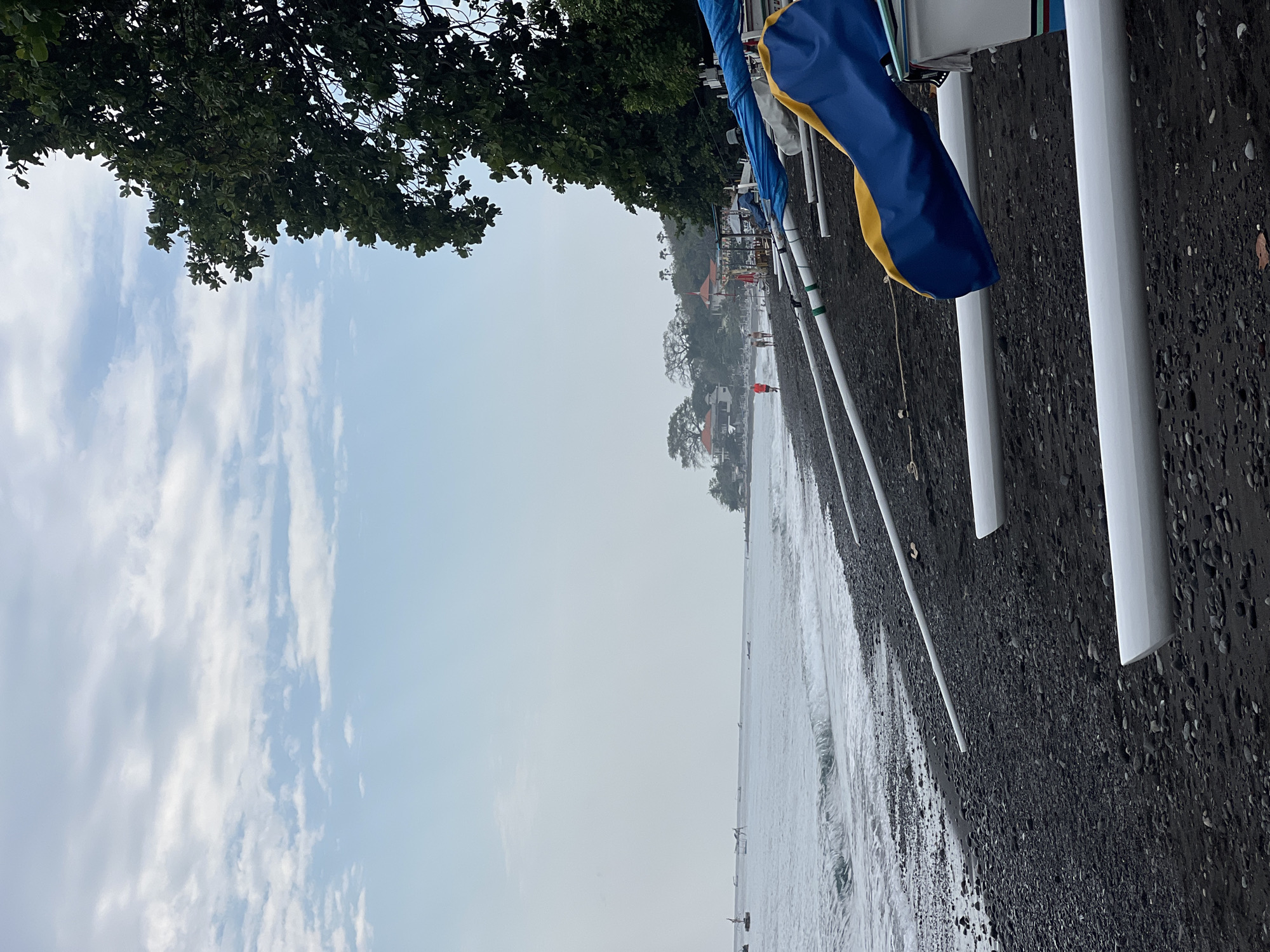 The image depicts a coastline in Abang, Indonesia, characterized by a black sand beach with numerous smooth pebbles. Several long, white outrigger canoe hulls with blue and yellow coverings are positioned on the shore. The ocean, with gentle waves, stretches to the horizon, where a hazy sky with scattered clouds is visible. In the distance, along the coast, a few buildings with red roofs can be seen, nestled amongst greenery and trees. Two figures are walking on the beach in the middle ground, one wearing a red shirt. Other small boats are anchored in the water further out. A dense green tree with a thick canopy occupies the right side of the frame, partially obscuring the sky.
