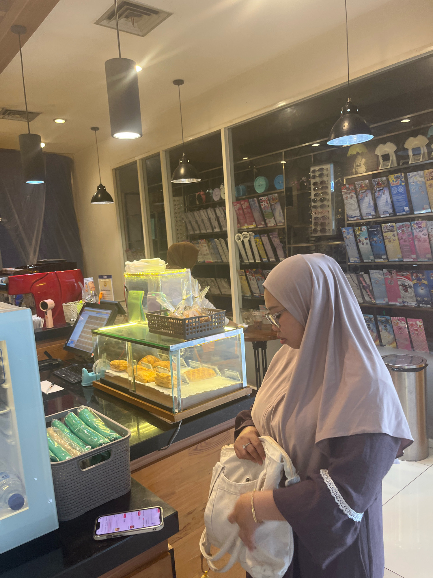 A woman wearing a light purple hijab and a dark purple abaya stands at a counter in what appears to be a cafe or bakery. She is looking down at a display case filled with pastries. In front of the display case is a cash register with a touchscreen monitor. To the left of the register, a grey woven basket contains several green wrapped items. A cell phone lies on the counter in front of the basket. Behind the woman, shelves are stocked with various items, including sunglasses and boxed products. The cafe has a modern interior with pendant lighting fixtures and a large window. The overall lighting suggests it is daytime.