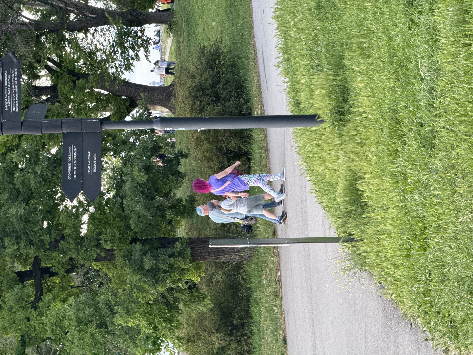 The image depicts a scene in Belgrade, Serbia, specifically in a park area. A metal signpost is visible, with directional signs pointing to "The Victor Monument" and "Roman Well" in both Serbian and English. The sign also identifies the "Institute for the Protection of Cultural Monuments of Belgrade." A paved walkway runs through the scene, flanked by grassy areas. Two individuals, a man and a woman with pink hair, are walking along the path.
