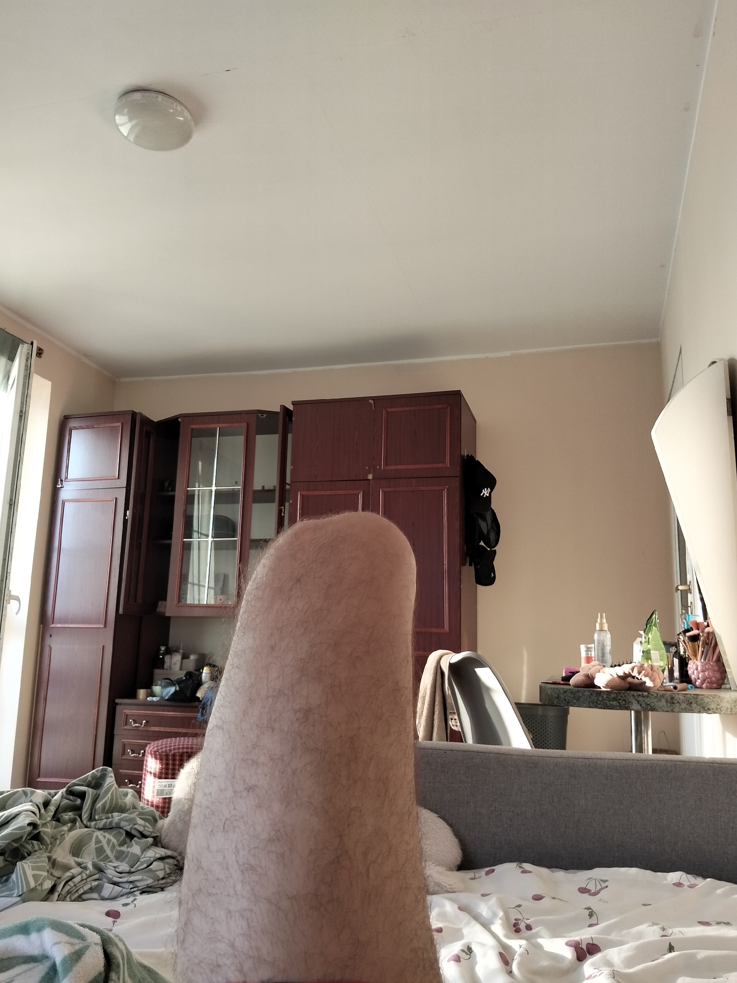 A person's hairy leg is the dominant subject in the foreground of this indoor scene, which appears to be a bedroom or living area. The leg is positioned vertically, taking up a significant portion of the lower and central frame. Behind the leg, a grey upholstered sofa is visible, covered with a white sheet patterned with cherries. Further into the background, a dark wooden wardrobe or cabinet unit with glass-fronted sections stands against a beige wall. A table with various items, including bottles, makeup brushes, and a pink pineapple-shaped holder, is situated to the right of the wardrobe. A window with a vertical blind is partially visible on the far left. The lighting suggests it is daytime. The scene does not provide any specific details to indicate the location as Brodnica, Poland.