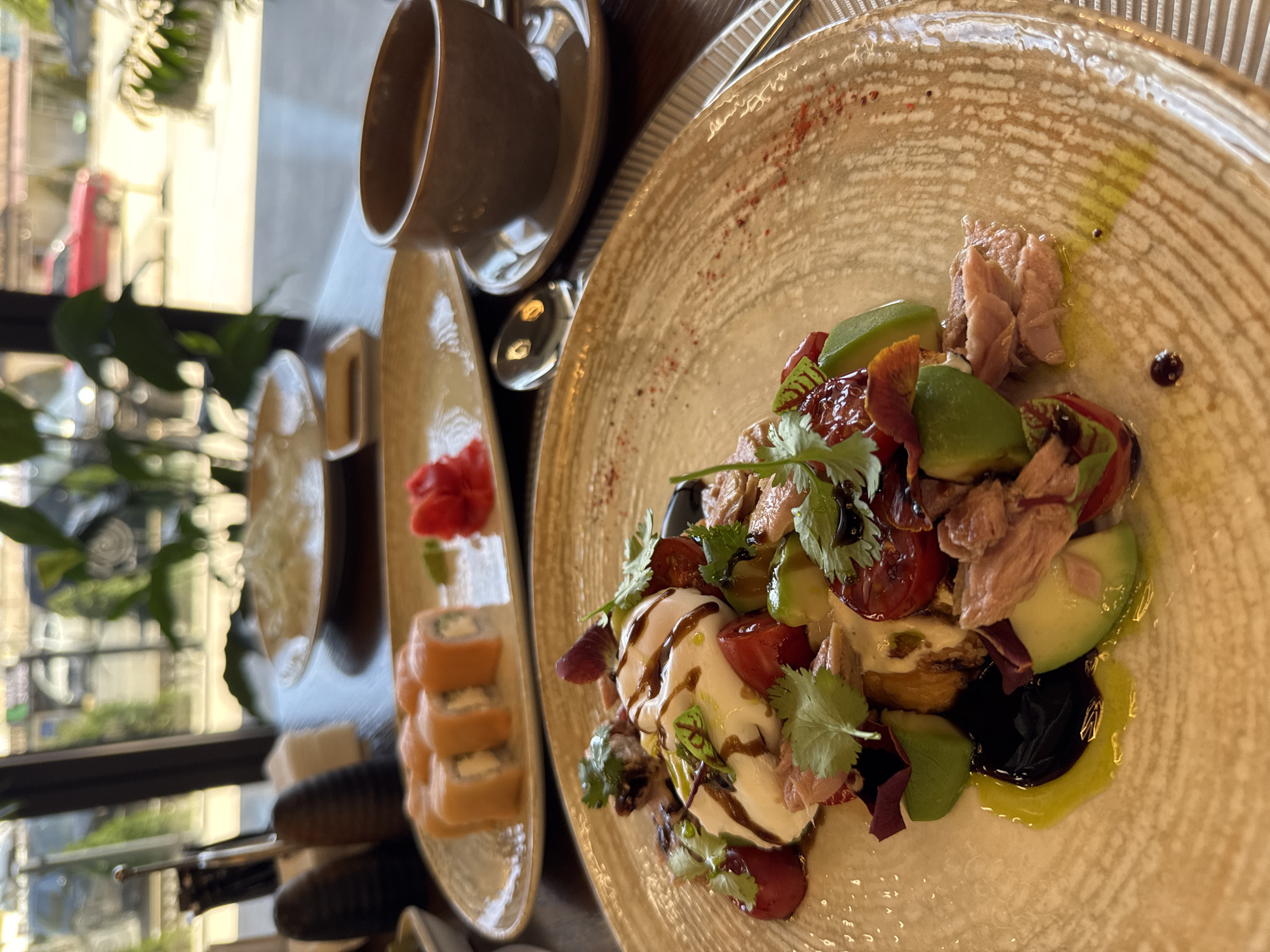 The image shows a table setting with food and drinks, likely in a restaurant. A plate with a salad, consisting of mozzarella, tomatoes, avocado, tuna, and balsamic glaze, sits on the right. Adjacent to it is a cup of coffee and a plate of sushi with ginger. The background includes a glimpse of the outside, possibly a window or open-air seating. The location is Çolpon-Ata, Kyrgyzstan.