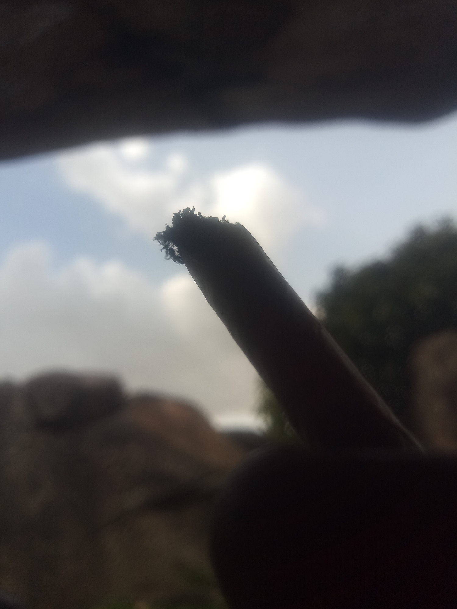 An extreme close-up shot shows the tip of a dark, cylindrical object angled upwards against a hazy sky. The object appears to be a rolled cigarette or similar item, with a frayed and uneven tip of ash. The background is a soft blur of light gray clouds and a hint of green foliage. The bottom of the frame is dominated by the dark, blurred shape of what appears to be a hand holding the object. The scene is set outdoors, with natural light suggesting daytime.