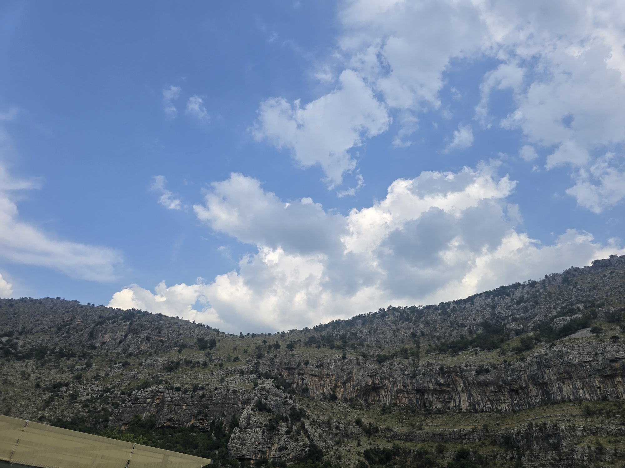 The image depicts a vast mountain landscape under a bright blue sky dotted with white cumulus clouds. The foreground on the left shows a portion of a slanted, light brown structure, possibly a roof or awning, with a textured surface. The main subject of the image is the rugged mountain range that dominates the middle and right portions. The mountains are steep, rocky, and appear to be composed of layers of stone with crevices and overhangs. Sparse green vegetation, including small trees and bushes, is scattered across the mountainside. The overall impression is a natural, undeveloped mountainous terrain. This view is likely of a mountainous region surrounding the city of Podgorica, Montenegro, given its geographical context. No people or specific actions are visible in the frame.