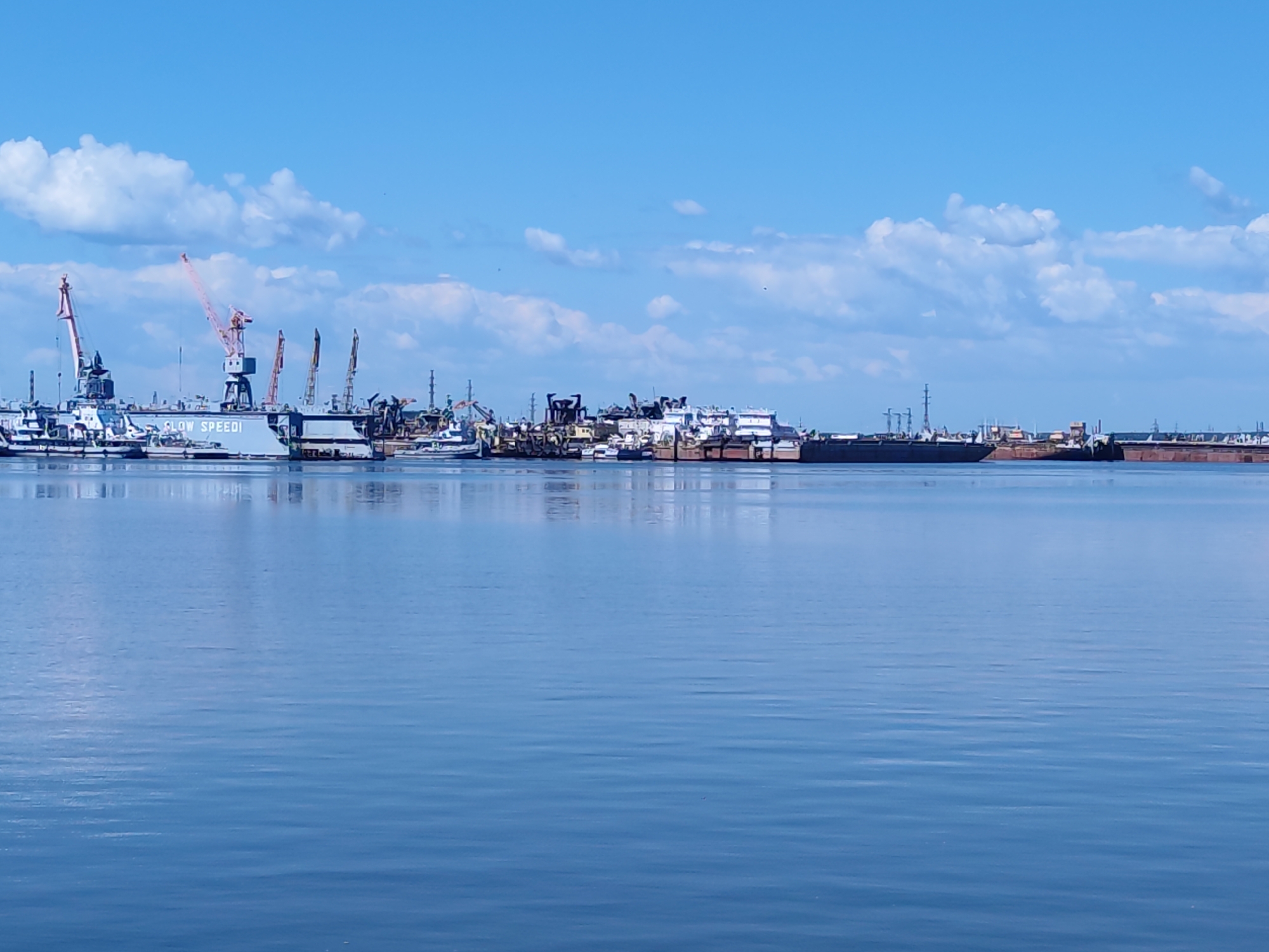 The image shows a panoramic view of a port in Chaikovsky, Russia, under a bright blue sky with scattered white clouds. The foreground is dominated by calm, blue water, reflecting the sky and the structures in the distance. Along the horizon, a series of industrial structures, including cranes of various sizes and colors, are visible. Several large vessels, possibly cargo ships or barges, are docked or moored in the port. One prominent structure appears to be a dry dock with a vessel inside. The overall scene depicts an active industrial port area.