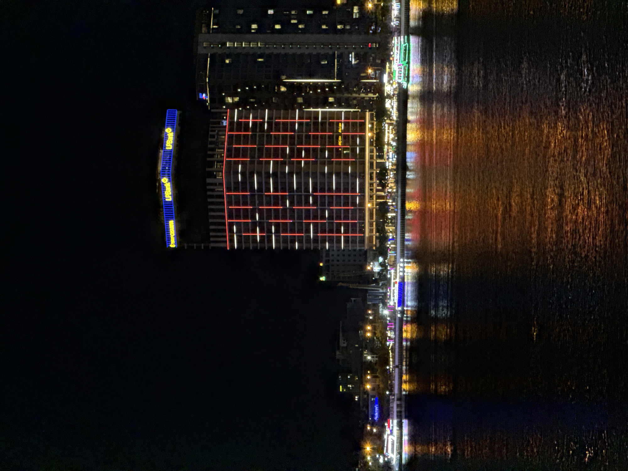 The image captures a nighttime cityscape in Đà Nẵng, Vietnam, viewed across a body of water, likely the Han River. A prominent, multi-story building dominates the mid-ground, featuring a distinctive blue-lit rooftop structure with several yellow "LPBank" logos. The facade of this building is adorned with vertical strips of red and white illumination. To its right, another tall building shows numerous illuminated windows. The distant shoreline reveals a sequence of smaller buildings and structures with various lights, suggesting an urban waterfront.
In the foreground, the river reflects the city's lights as blurred streaks of orange, yellow, red, blue, and white across its subtly rippled surface. Two illuminated vessels are visible near the far bank. One boat, positioned to the left, exhibits bright blue lighting. Another boat, further to the right, is prominently illuminated in green. A brightly lit promenade or street with streetlights runs along the river's edge on the far bank. The sky above is uniformly dark, and no discernible individuals or specific actions are visible within the frame.