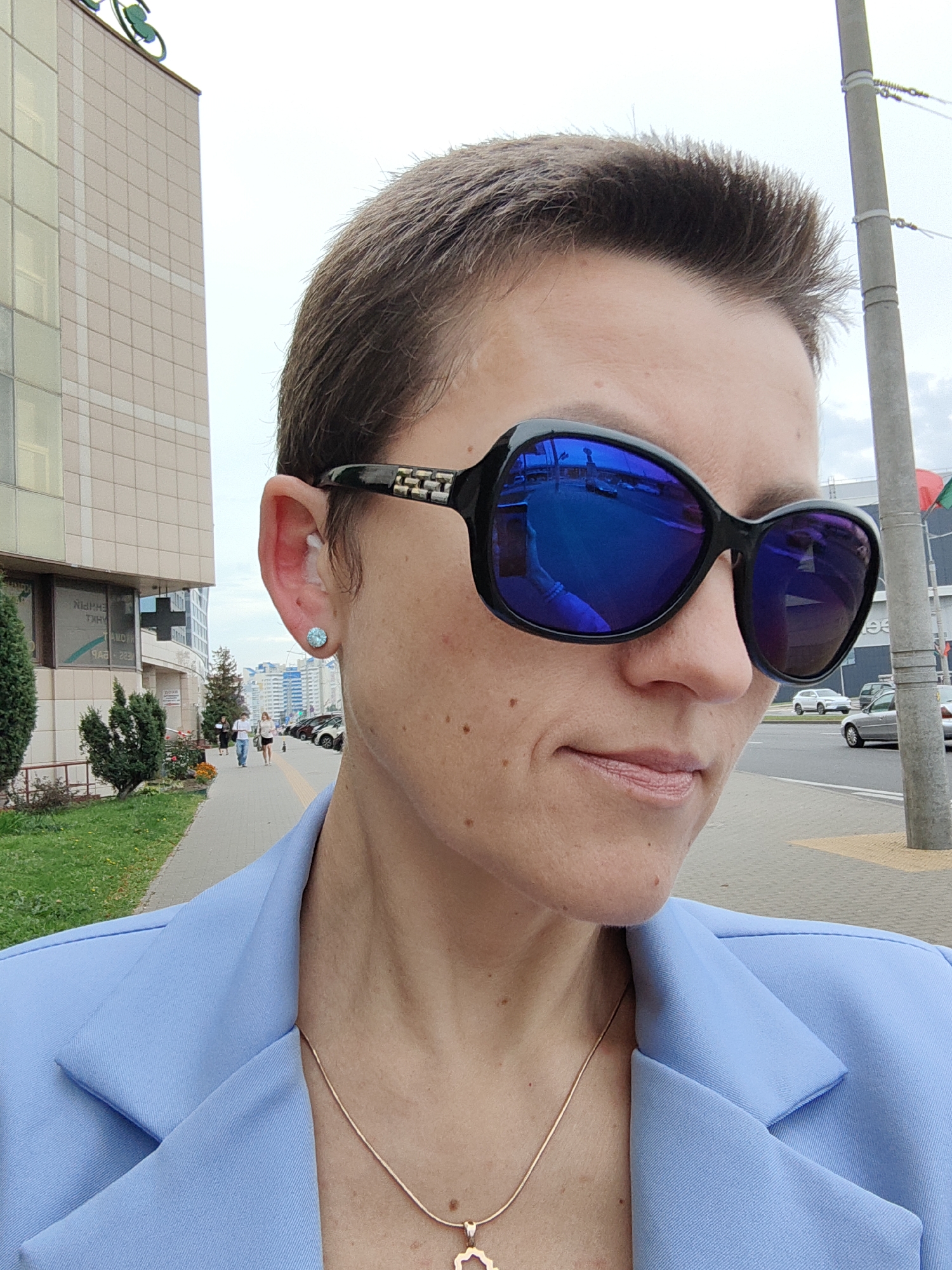 A medium shot captures a woman with short brown hair wearing black sunglasses and a light blue blazer. She is standing on a sidewalk in Minsk, Belarus, with a modern building to her left and a street with cars to her right. A concrete pole stands to her right. The woman is looking slightly to her right, with a faint smile. Her face is visible from the nose up. Her ear, adorned with a small stud earring, is also visible. A thin gold necklace with a pendant rests on her blazer. In the background, people are walking on the sidewalk, and trees and other buildings are visible. The sky is overcast.