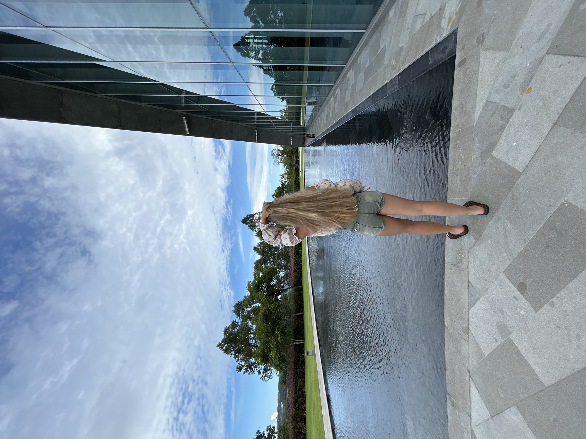 The image captures an outdoor scene featuring a person standing beside a long, tranquil reflecting pool. The person, seen from behind, has long blonde hair and is wearing a light-colored, floral patterned top and olive green shorts. Their hands are raised to the back of their head.
To the left, a modern building with large, reflective glass panels mirrors the bright blue sky with white clouds and the surrounding trees. The reflecting pool, dark and still, also clearly mirrors the sky and distant greenery, creating a sense of depth and openness. A grey paved walkway with geometric patterns is visible to the right of the person and the pool. Beyond the pool, a strip of vibrant green lawn is bordered by a dense line of trees and shrubs, some adorned with small pink or purple blossoms. The sky above is partially cloudy, indicating bright daylight. The overall setting suggests a well-maintained, contemporary landscape, possibly in an architectural park, resort, or public space in Kuta Selatan, Indonesia. There is no visible text in the image.