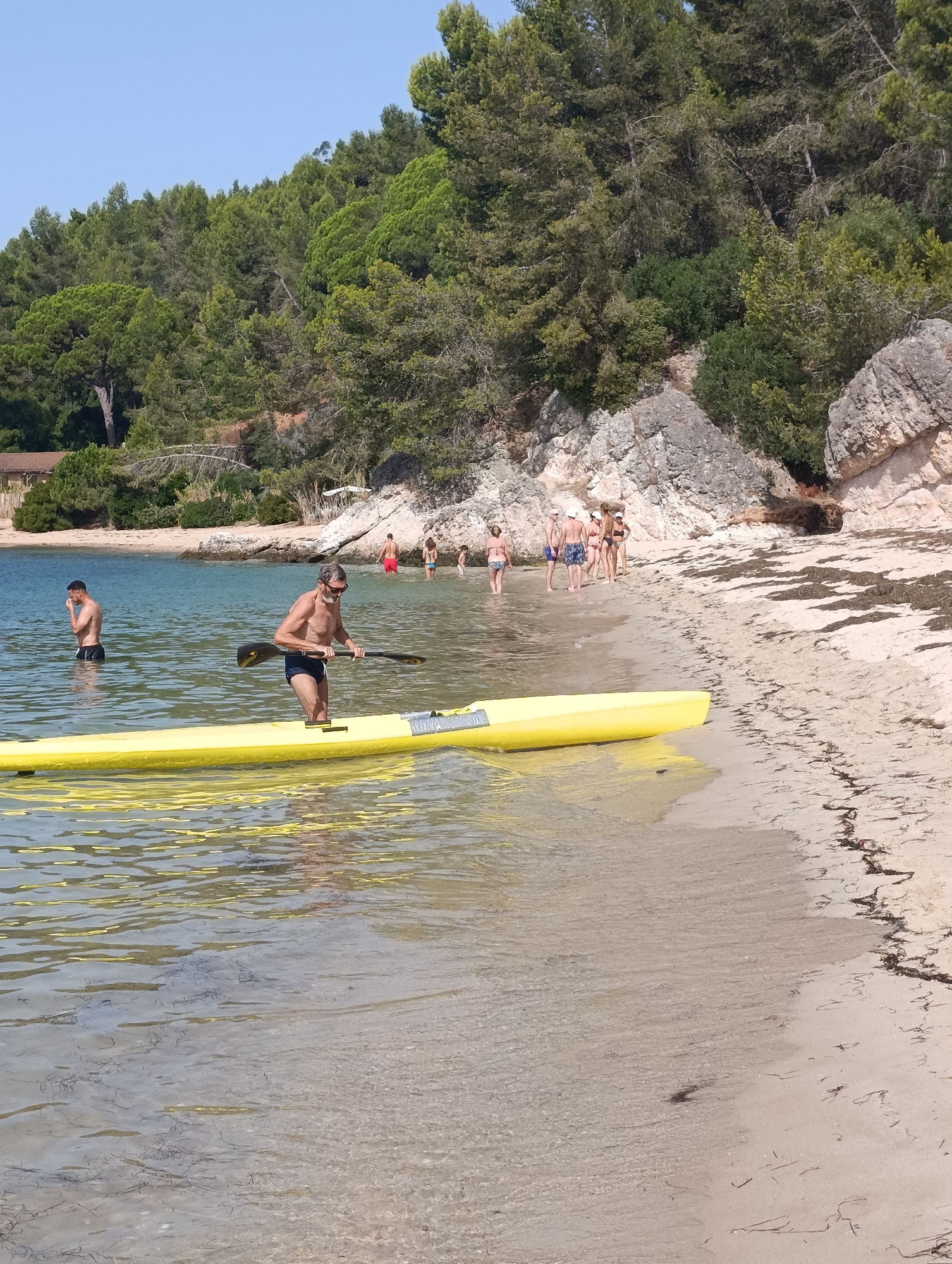 The image shows a sunny day at a beach with clear, calm water. A man in dark blue swim trunks and sunglasses is standing in shallow water next to a bright yellow paddleboard, holding a paddle. Another man in dark swim trunks stands further in the water to the left, with his hand near his face. In the background, a group of people are standing on the sandy beach and in the water. The beach is backed by a rocky, tree-covered hillside. The scene appears to be a natural coastal environment, but no specific landmarks in Setúbal, Portugal, are identifiable.