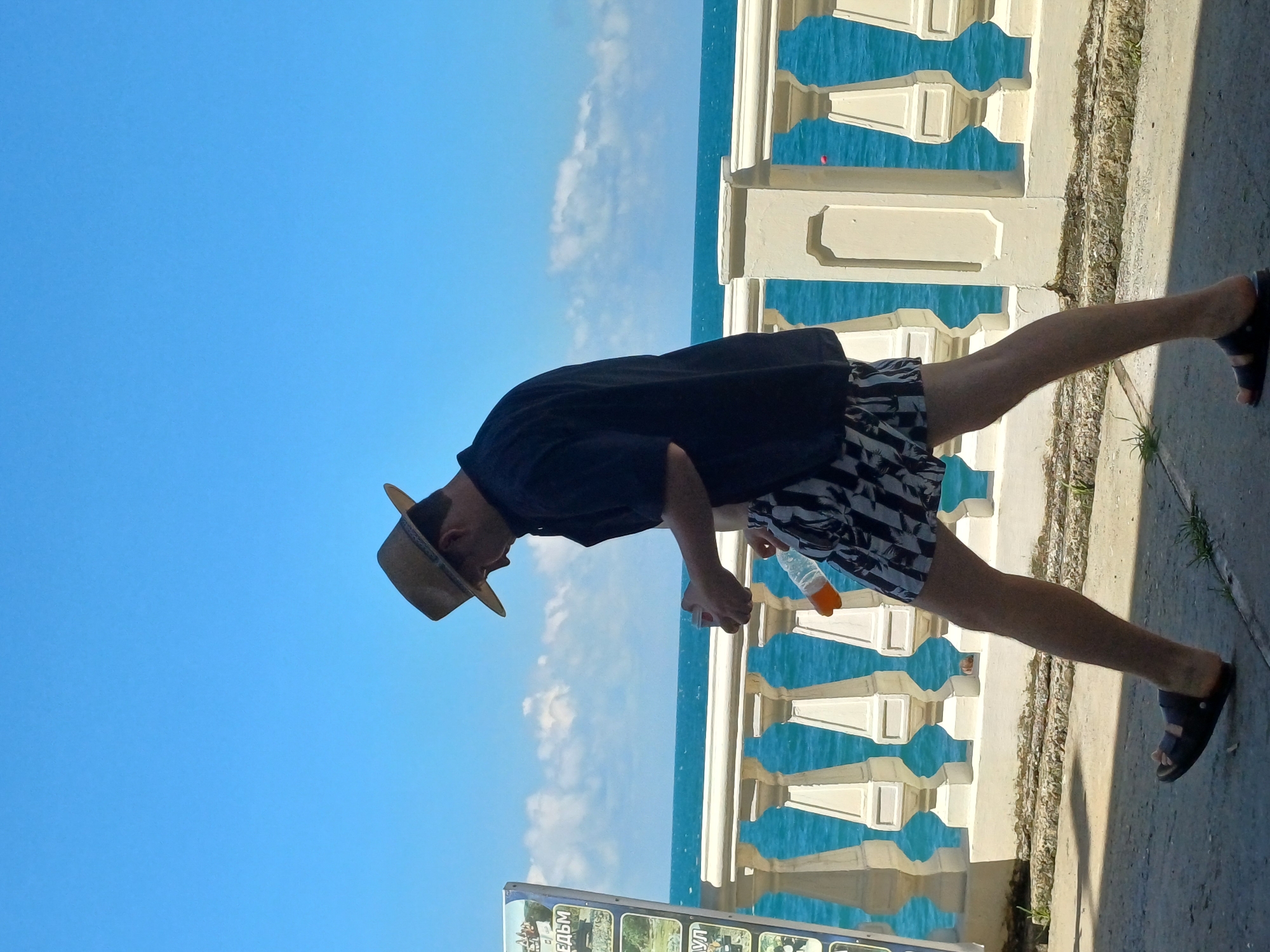The image shows a person wearing a black shirt, patterned shorts, and a straw hat, standing on a paved walkway next to a white balustrade. The person is holding a clear plastic bottle with an orange cap. The sea is visible behind the balustrade, with a bright blue sky and some clouds above. The sun is casting a shadow on the walkway, suggesting it is daytime. The overall scene depicts a person enjoying a day outdoors by the sea, possibly in a resort area. There is some visible text on a sign at the bottom of the image, which appears to be in Russian, with words like "ВЕДЬМ" and "КУЛЬТУРЫ". The location context suggests this might be in Sochi, Russia, a coastal city known for its beaches and resorts. The person's posture and attire suggest a relaxed, leisurely atmosphere.