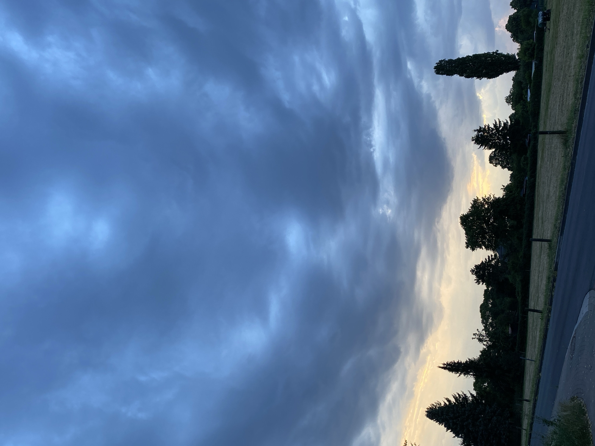 The image depicts a cloudy sky over a rural landscape, likely on the outskirts of Rinteln, Germany. The sky is dominated by heavy, dark blue and gray clouds, with lighter patches suggesting a potential break in the clouds or sunset light. Below the clouds, a line of dark green trees forms a silhouette against the horizon. The trees vary in shape and size, with some appearing to be evergreen conifers and others more deciduous. In the foreground, a paved road curves to the left, bordered by a grassy field. The field is dry and appears to have been recently mown, with a few wooden posts marking a boundary. There are no people or animals visible in the image. The overall impression is one of a tranquil, slightly overcast rural scene.